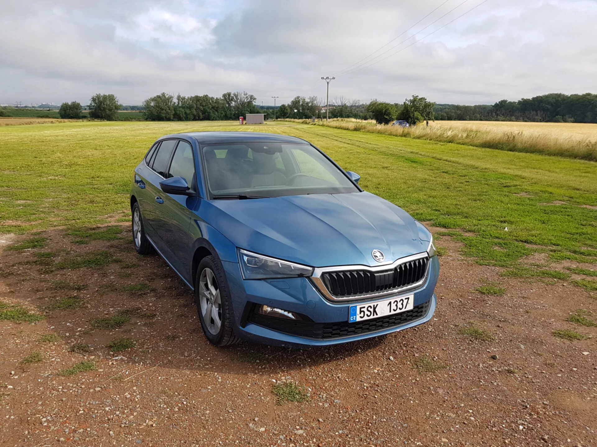 Skoda Scala azul 2019 visto desde un ángulo frontal de tres cuartos destacando diseño, faros LED, parrilla cromada, ruedas de aleación de múltiples radios, señales de giro integradas en los espejos laterales y ventanas tintadas, colocado sobre grava con un campo en el fondo bajo cielos nublados.