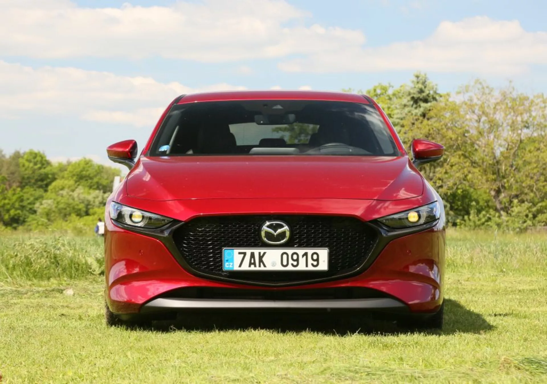 Vehículo Mazda rojo con faros LED y parrilla cromada estacionado en el césped bajo un cielo despejado, mostrando el diseño deportivo del frontal y la estética del vehículo.