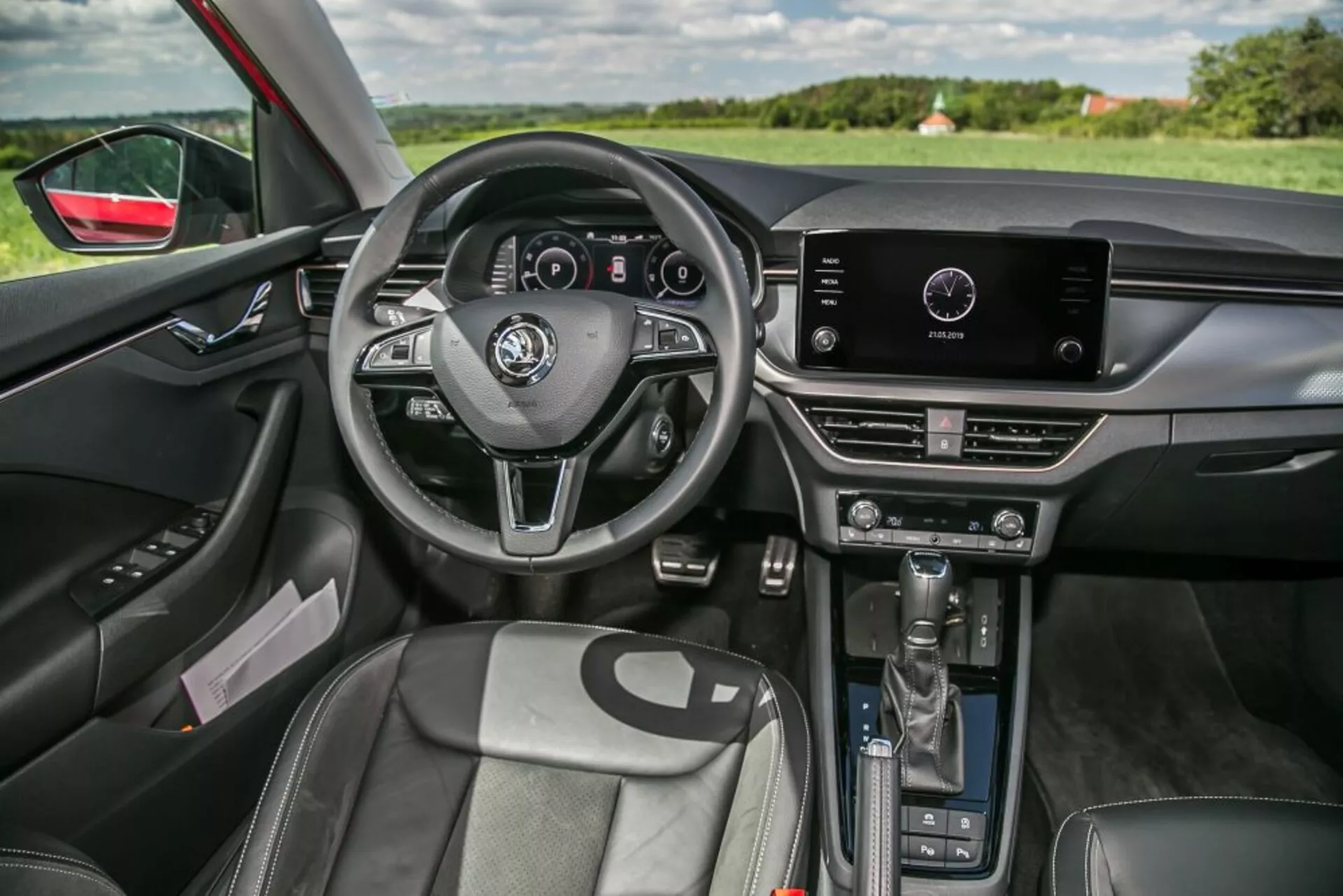 Vista interior del Skoda Scala 2019 desde el lado del pasajero delantero con volante de tres radios con controles integrados y cuadro de instrumentos digital, sistema de infoentretenimiento táctil en la parte superior de la consola central con controles manuales del climatizador debajo, palanca de cambios automática, asientos delanteros reforzados con tapicería similar al cuero, panel de puerta del conductor con ajustes eléctricos de ventanas y espejos, iluminación ambiental interior, espejo retrovisor con atenuación automática y visibilidad del paisaje a través del parabrisas y ventana del lado del conductor, destacando el diseño ergonómico y la tecnología integrada.
