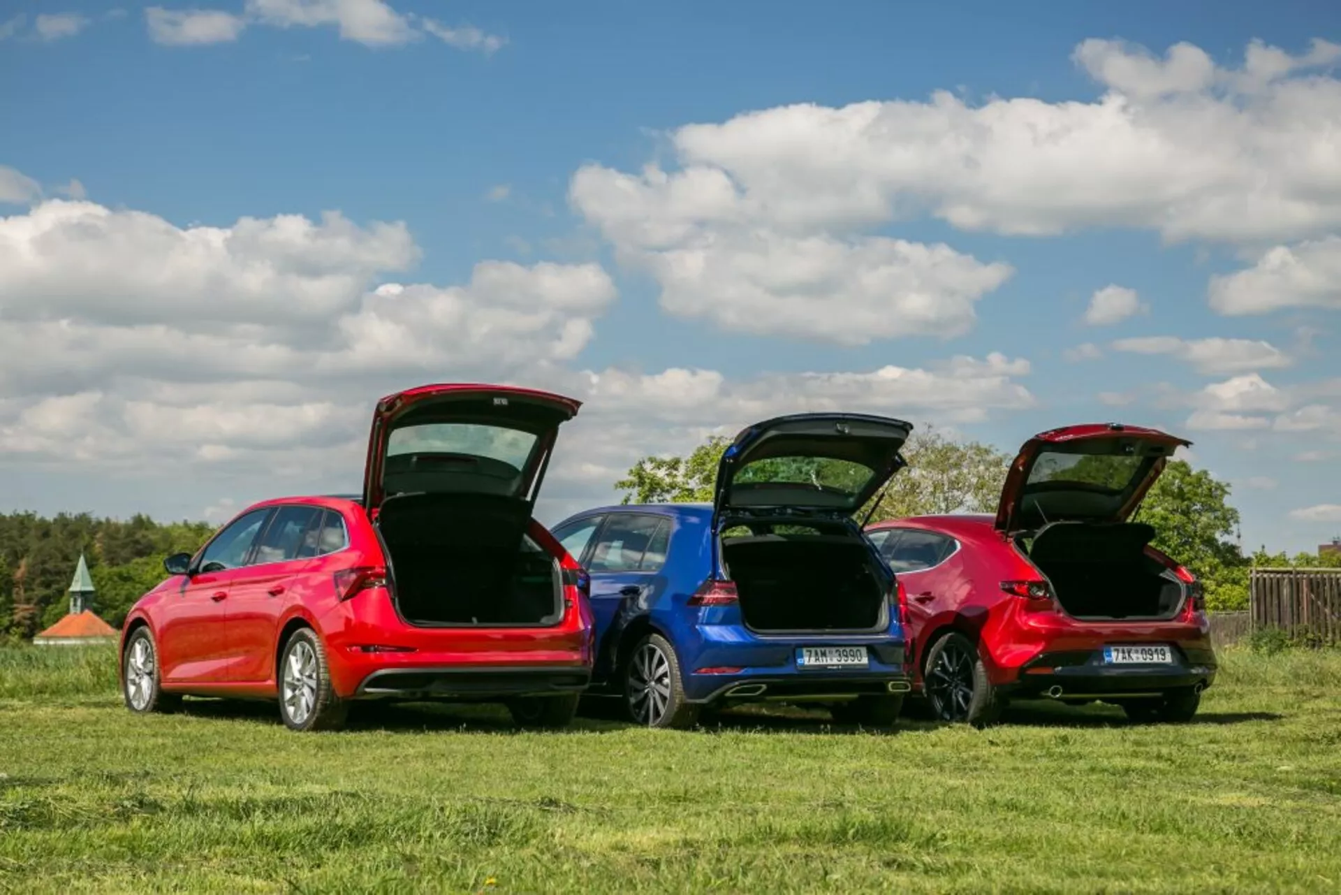 Modelos de Skoda Scala 2019 en rojo y azul estacionados en un campo de hierba, mostrando perfiles traseros, maleteros abiertos para funcionalidad de espacio de carga y detalles de diseño bajo cielos despejados.