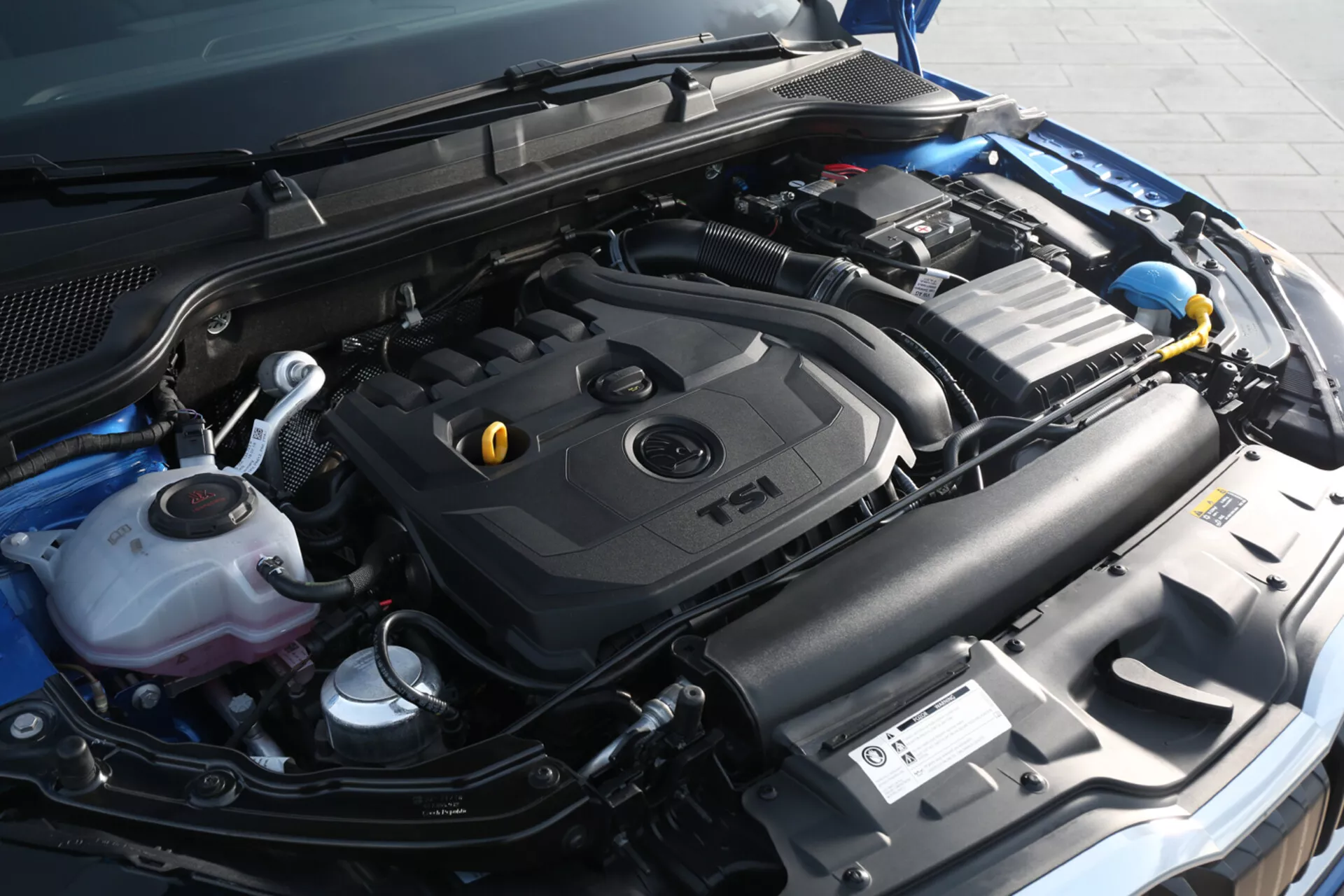 Vista detallada del compartimento del motor de un Skoda Scala 2019, destacando la cubierta del motor turboalimentado TSI y componentes incluyendo el depósito de refrigerante, contenedor de líquido limpiaparabrisas, batería del coche y sistema de admisión desde un ángulo frontal ligeramente a la derecha