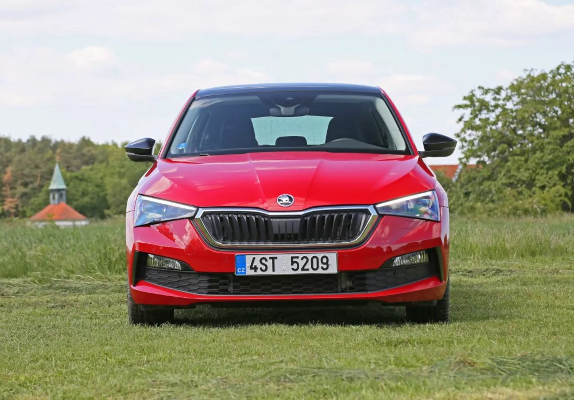 Skoda Scala 2019 en rojo situado en un campo herboso con vista frontal, mostrando la parrilla frontal del coche, faros afilados y el logo de Skoda, contra un fondo natural con árboles y un toque de cielo azul.