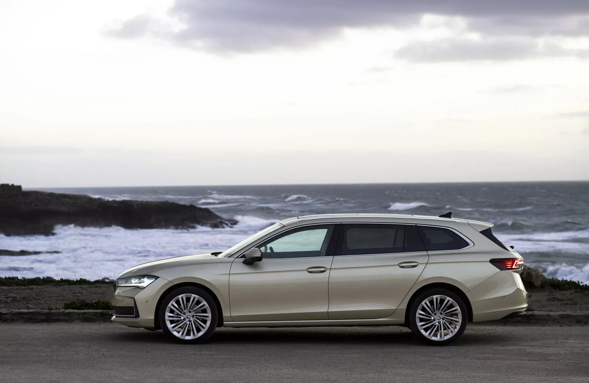 Familiar de color claro Skoda Superb Combi 2024 estacionado en una carretera costera con vista al océano, con una línea de techo inclinada, cinco puertas, ruedas grandes de aleación con múltiples radios, ribetes de ventana cromados y espejos laterales del color de la carrocería contra un fondo de olas rompiendo y una luz cálida al atardecer.