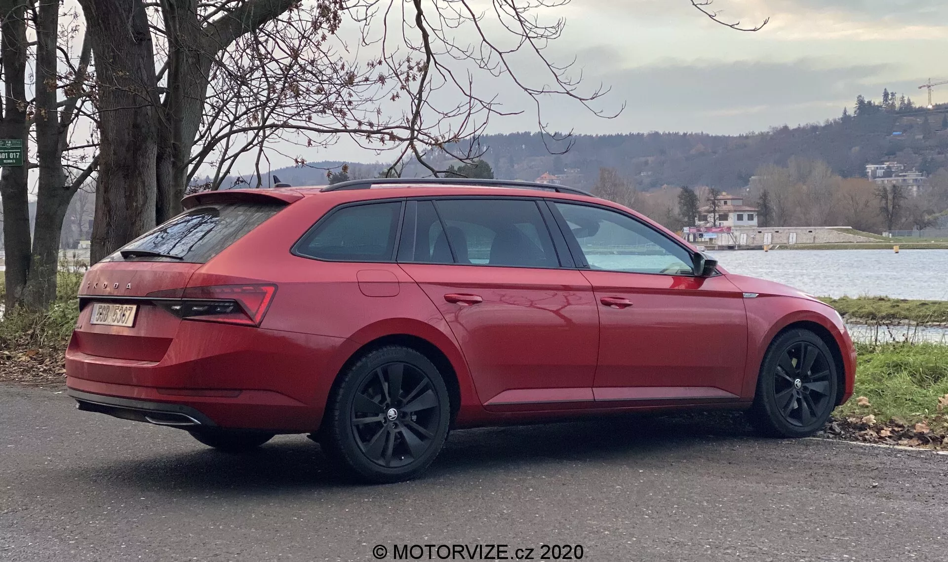 Un coche familiar Skoda Superb 2019 rojo estacionado al aire libre, visto en ángulo trasero de tres cuartos mostrando el lado derecho del coche. Los elementos de diseño clave incluyen grandes llantas de aleación oscuras, ventanas traseras y laterales inclinadas, luces traseras envolventes, portón trasero cerrado con insignia, parachoques trasero con difusor y doble escape, rieles de techo y espejo lateral exterior a juego con el color. El entorno presenta vegetación y un cuerpo de agua en el fondo, sugiriendo un entorno suburbano o semirrural tranquilo.