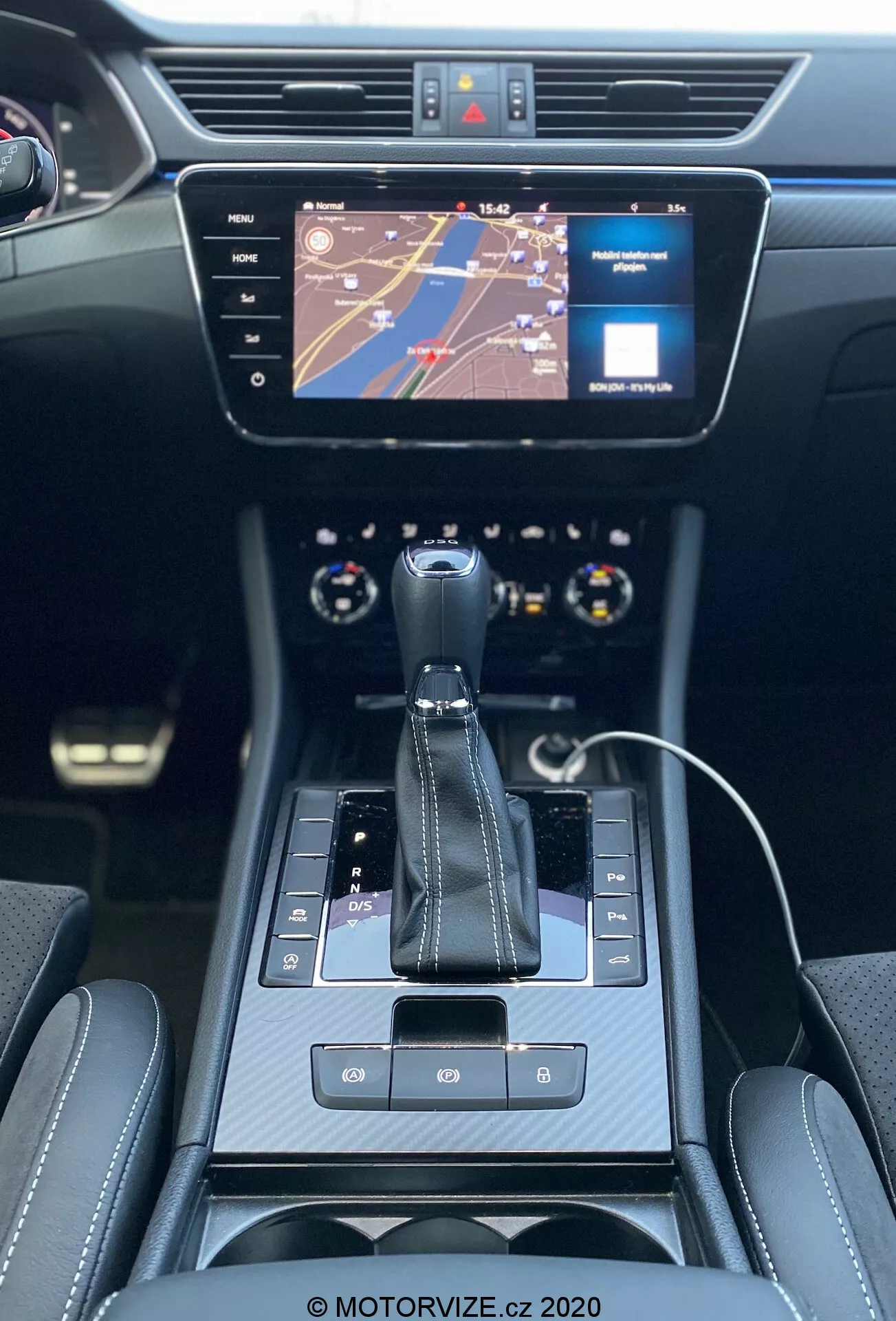 Vista interior del Skoda Superb Limousine 2019, enfocada en la consola central y el tablero de instrumentos desde la perspectiva del asiento trasero. Presenta una palanca de cambios automática con 'P' visible para estacionamiento, botón de freno de mano electrónico, botones táctiles y una interfaz táctil. Una pantalla táctil de infoentretenimiento muestra un mapa y un indicador de medios. Controles de clima de doble zona debajo de la pantalla, flanqueados por rejillas de ventilación. Interior moderno y elegante en negro con detalles cromados y asientos lujosos con costuras visibles. Parte superior del volante multifuncional y parte del cuadro de instrumentos con diales analógicos visibles. Cable de carga de smartphone indicando conectividad.