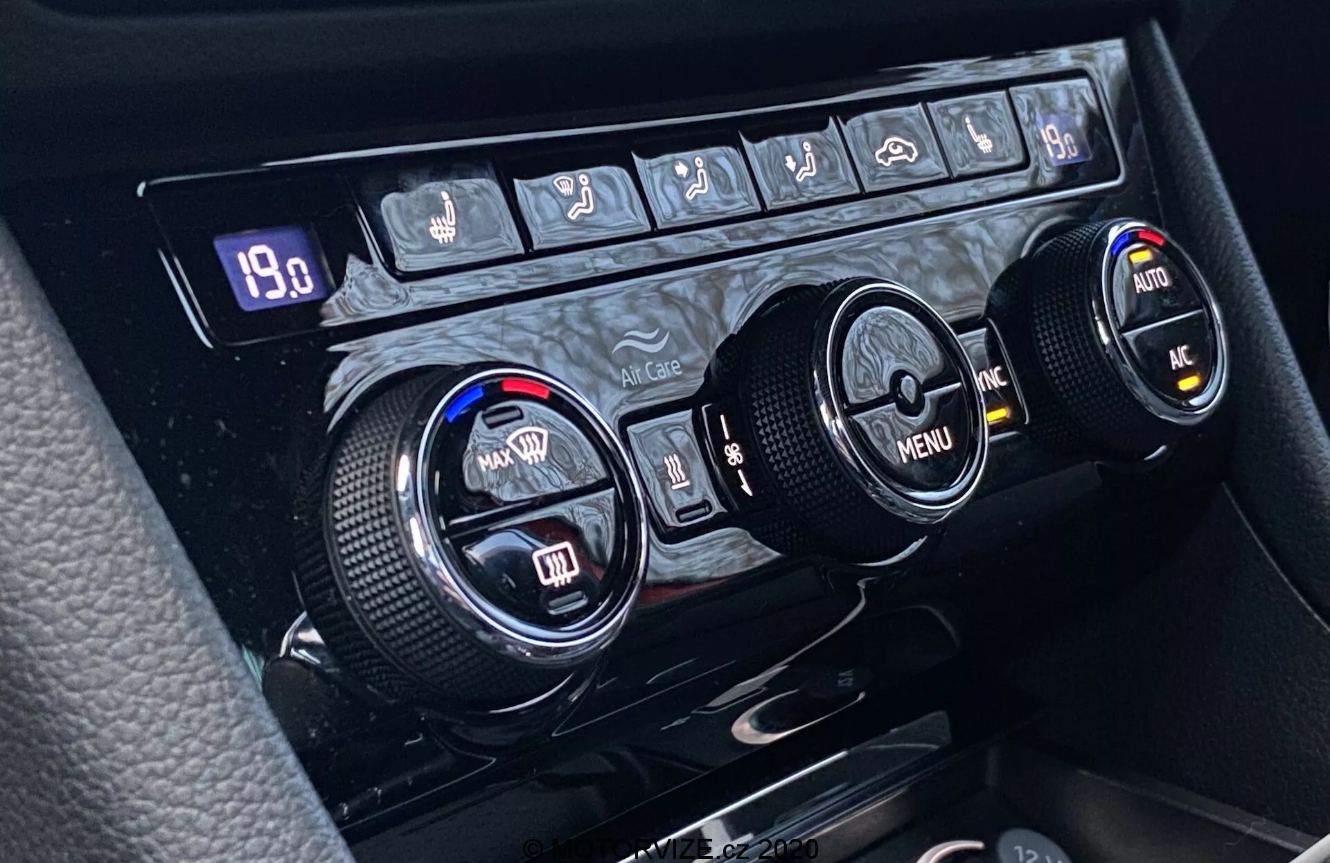 Primer plano del sistema de control de clima del Škoda Superb 2019 situado en la consola central, con tres esferas circulares para el control de temperatura, la navegación por el menú y funciones adicionales. La esfera izquierda ajusta la temperatura para el conductor y la derecha para el pasajero, mientras que la esfera central ofrece navegación por el menú con flechas direccionales. Los bordes texturizados aseguran un uso ergonómico. Sobre las esferas, una fila de botones incluye controles para el desempañado de la ventana trasera, el desempañado de la ventana delantera, la circulación del aire, el control climático automático, el aire acondicionado y posiblemente una función de emergencia. Una pantalla digital muestra '19.0', indicando la temperatura configurada o un valor relacionado.