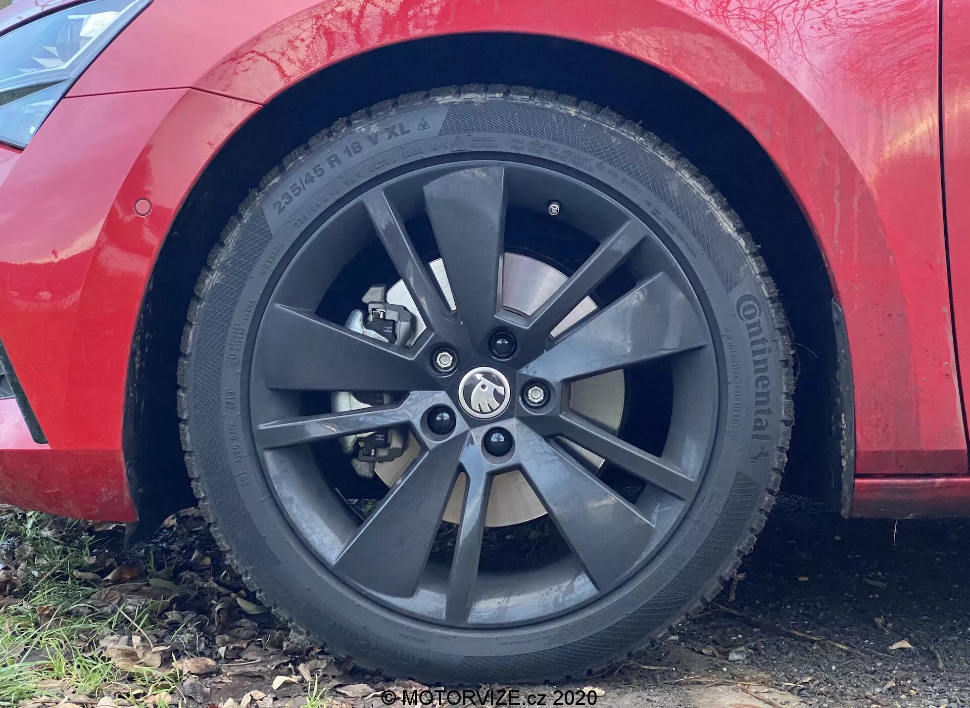 Vista en primer plano de la rueda delantera del lado del pasajero y el guardabarros del Skoda Superb 2019. La imagen muestra un diseño de rueda moderno, de múltiples radios, con un acabado oscuro en grafito o negro, convergiendo en el cubo con un emblema de coche no identificable. El neumático parece ser de la marca Continental, con especificaciones visibles pero no completamente legibles. La iluminación sugiere un entorno diurno, con la pintura roja limpia del coche apenas visible.