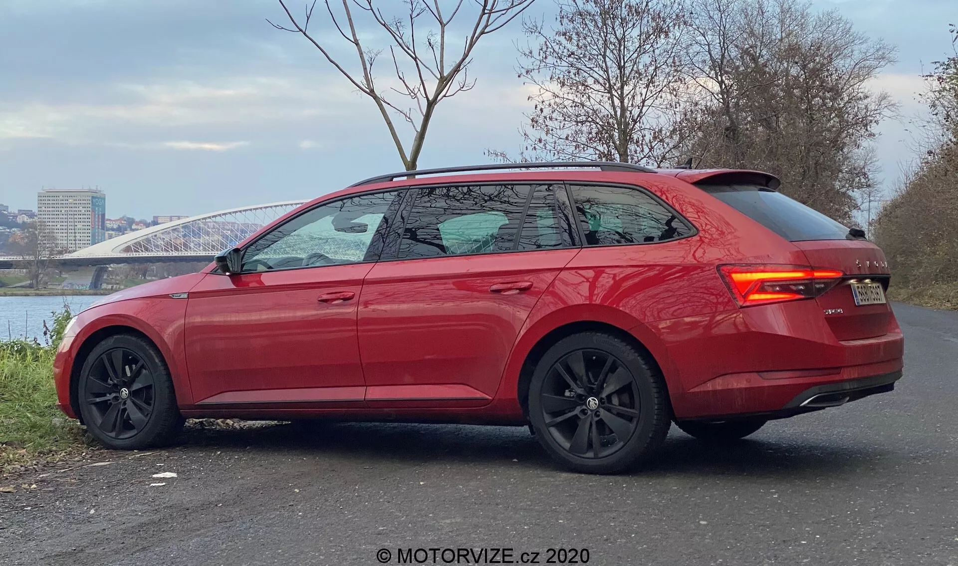 La imagen presenta un Skoda Superb 2019 moderno y vibrante de color rojo, visto desde un ángulo de tres cuartos trasero. Estacionado en una zona pavimentada con un río y un puente en el fondo, el coche muestra líneas elegantes, un techo trasero inclinado, ventanas ligeramente tintadas con adornos cromados y un alerón trasero integrado. El diseño deportivo se destaca por grandes ruedas de aleación negras con pinzas de freno visibles, espejos laterales negros contrastantes, manijas de puertas al ras, luces traseras LED y sensores de estacionamiento en el parachoques trasero, indicando un alto nivel de acabado con características de rendimiento y asistencia al conductor avanzadas.