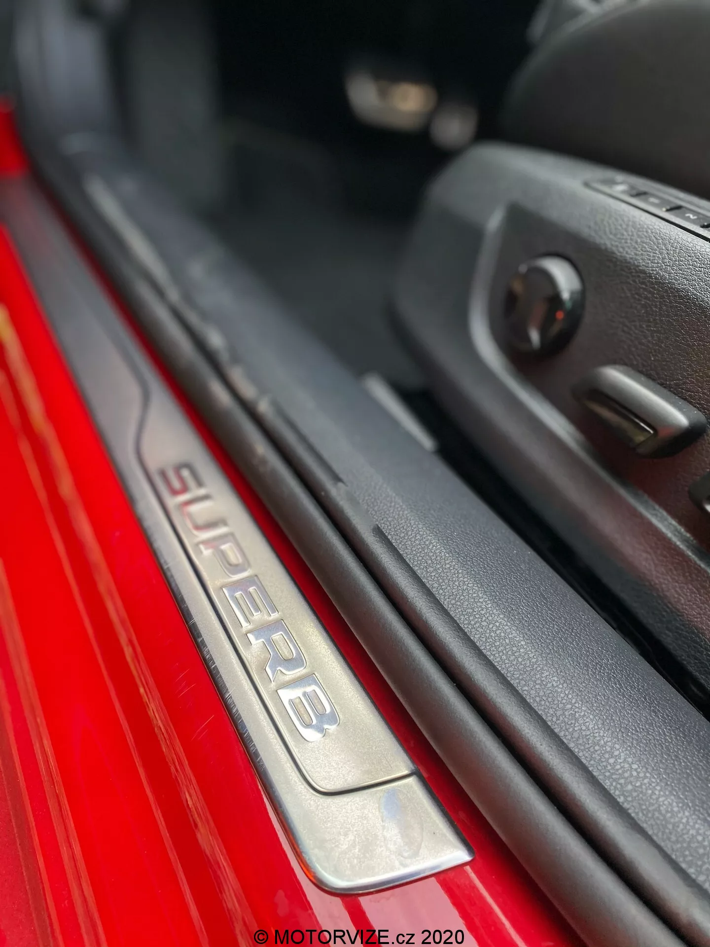 Vista de cerca de la placa del umbral de la puerta del lado del pasajero del Skoda Superb 2019. La imagen presenta un ángulo inclinado que muestra la inscripción 'SUPERB' en la placa de umbral de acero inoxidable cepillado, iluminada para un atractivo estético mejorado. La pintura exterior roja contrasta bruscamente con el interior oscuro. Se puede ver el borde de la puerta con el panel de plástico y los controles de las ventanas eléctricas, resaltando características avanzadas. La poca profundidad de campo destaca la placa del umbral, creando un efecto bokeh en el fondo.