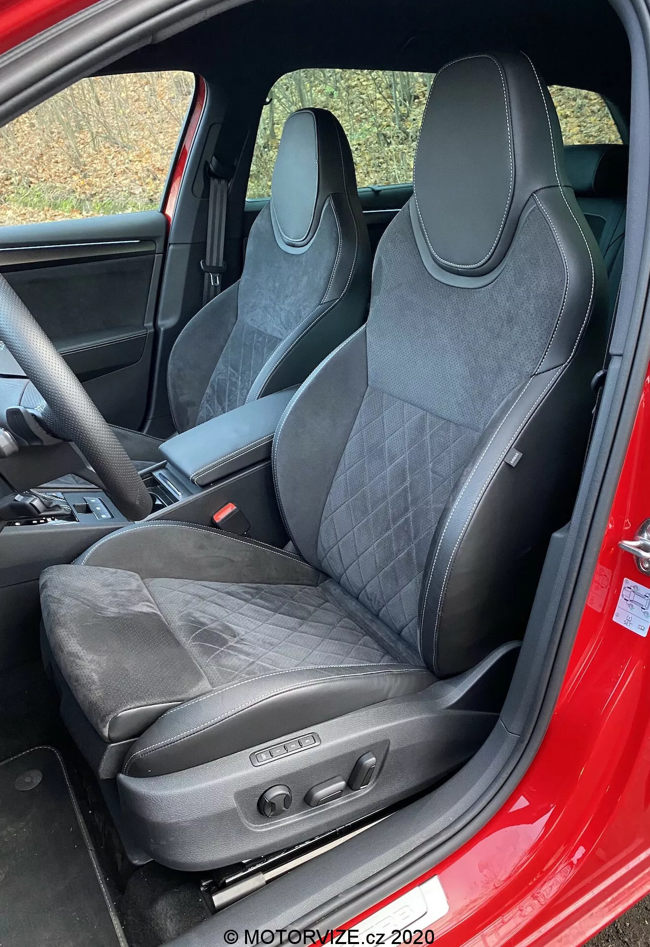 Interior del Škoda Superb 2019 centrado en el asiento del conductor. El asiento de estilo deportivo con tapicería de cuero y material similar a ante, costuras acolchadas, reposacabezas integrado, contornos pronunciados en los hombros y controles de ajuste electrónico indica un diseño orientado al rendimiento, lujo y características avanzadas de confort. El marco de la puerta visible de color rojo sugiere que el exterior podría ser rojo.