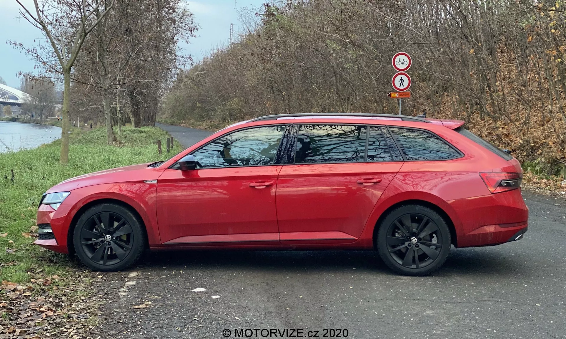 Imagen de perfil lateral de un moderno coche familiar Skoda Superb 2019 (Superb-Limousine) en rojo, mostrando su línea de techo alargada, llantas de aleación negras y espejos laterales del color de la carrocería con intermitentes integrados. El coche está aparcado en un camino de asfalto junto a una zona con césped, con árboles y un cuerpo de agua al fondo, bajo la luz natural del día.