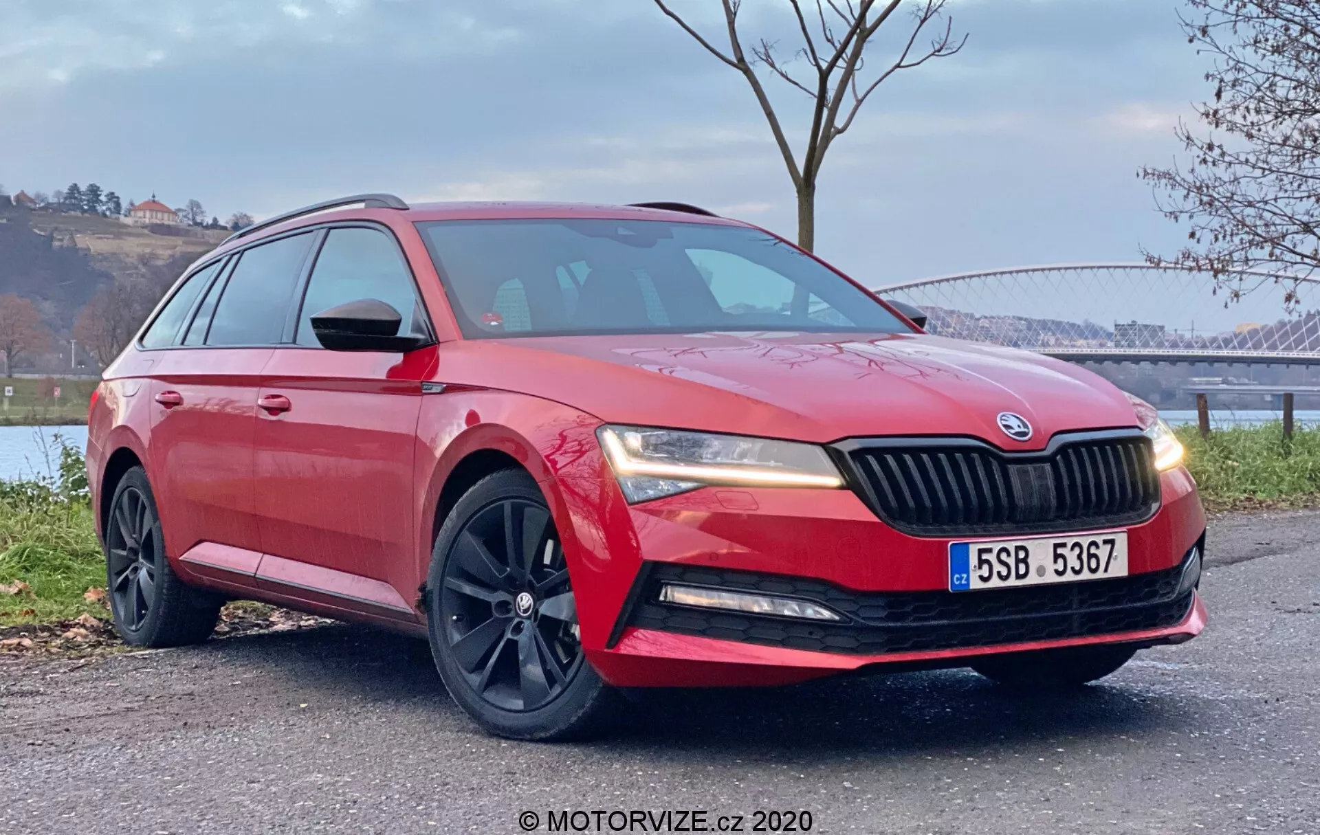 Vista de un cuarto delantero a baja altura del Škoda Superb 2019 rojo estacionado al aire libre, mostrando las líneas de diseño, parrilla, ruedas, perfil lateral y las aleaciones oscuras deportivas de la wagon frente a un paisaje pintoresco con agua y árboles.