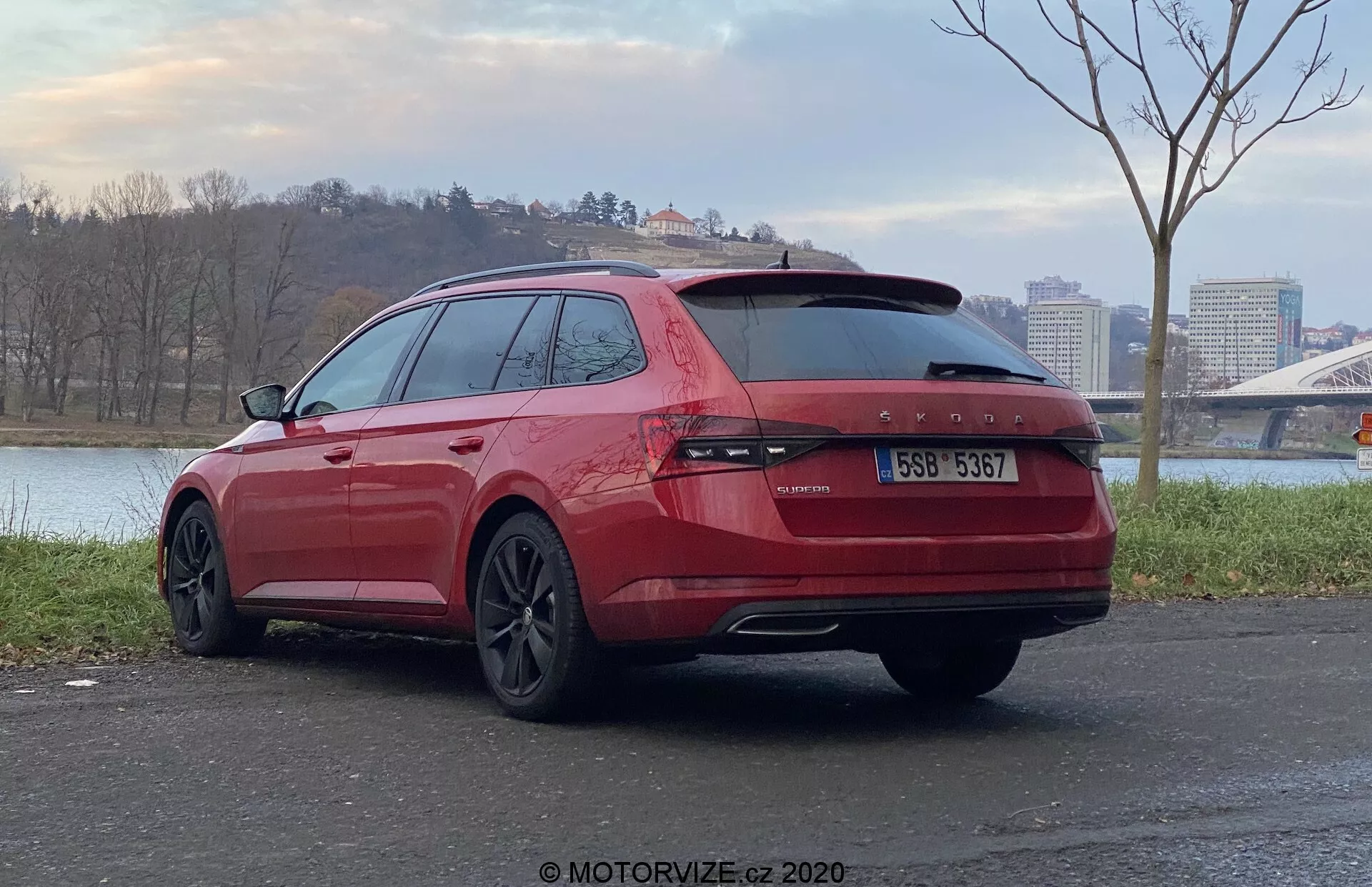 Vista trasera en ángulo de una camioneta Skoda Superb 2019 roja estacionada al aire libre, enfocándose en la parte trasera y lateral del pasajero del vehículo. Con luces traseras continuas a lo largo del portón, dos tubos de escape, vidrios polarizados, ruedas de aleación con acabado oscuro y ubicada en una superficie pavimentada con un cuerpo de agua y un puente en el fondo.