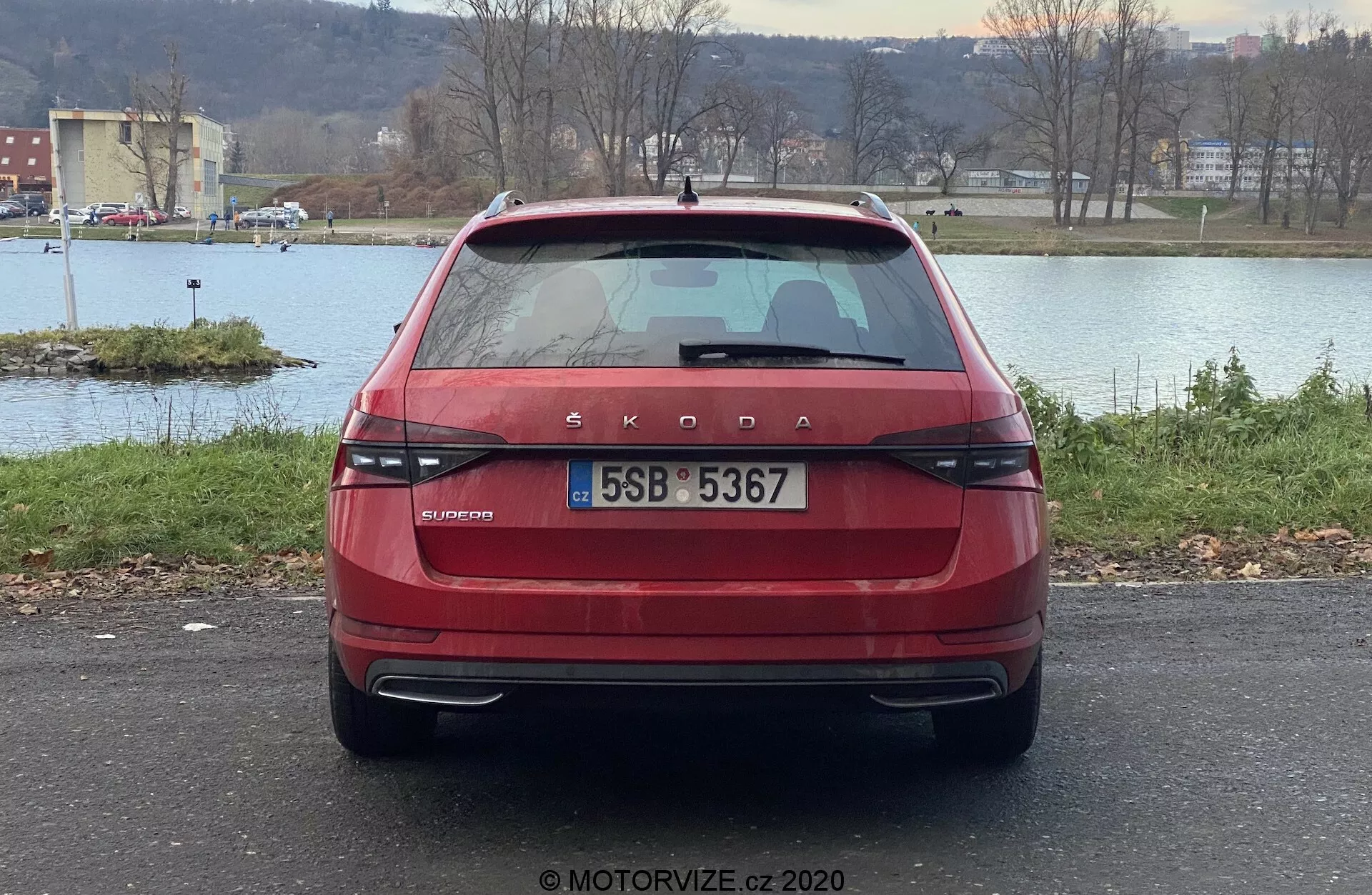 Vista trasera de un Škoda Superb 2019 rojo limusina, mostrando la distintiva marca Škoda y la inscripción del modelo OCTAVIA en el maletero. La imagen resalta las modernas luces traseras alineadas horizontalmente, la matrícula centrada, el limpiaparabrisas trasero, la ventana trasera tintada con líneas de desempañado, la luz de freno montada en alto, los sensores de aparcamiento en el parachoques y los reflectores. El coche está aparcado en exteriores con un cuerpo de agua, árboles y un horizonte en el fondo.
