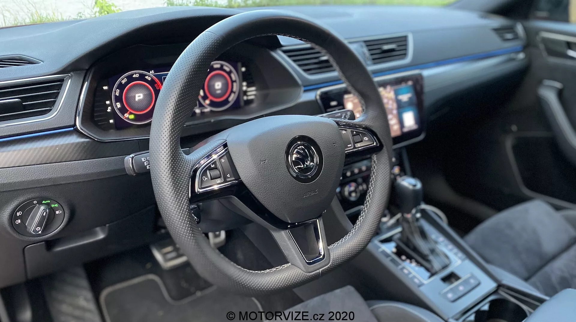 Vista interior de un Skoda Superb Limousine 2019 destacando el área de la cabina del conductor. Volante de tres radios central con el logotipo del fabricante y controles para sistema de audio y de a bordo, incluyendo levas de cambio. Paneles de instrumentos digitales detrás, indicando velocímetro y tacómetro. Pantalla táctil de infoentretenimiento inclinada hacia el conductor, control de clima de doble zona, palanca de cambios deportiva, selector de modo de conducción y asientos de cuero de calidad con costuras acentuadas sugieren un vehículo moderno y de alta gama con tecnología avanzada.