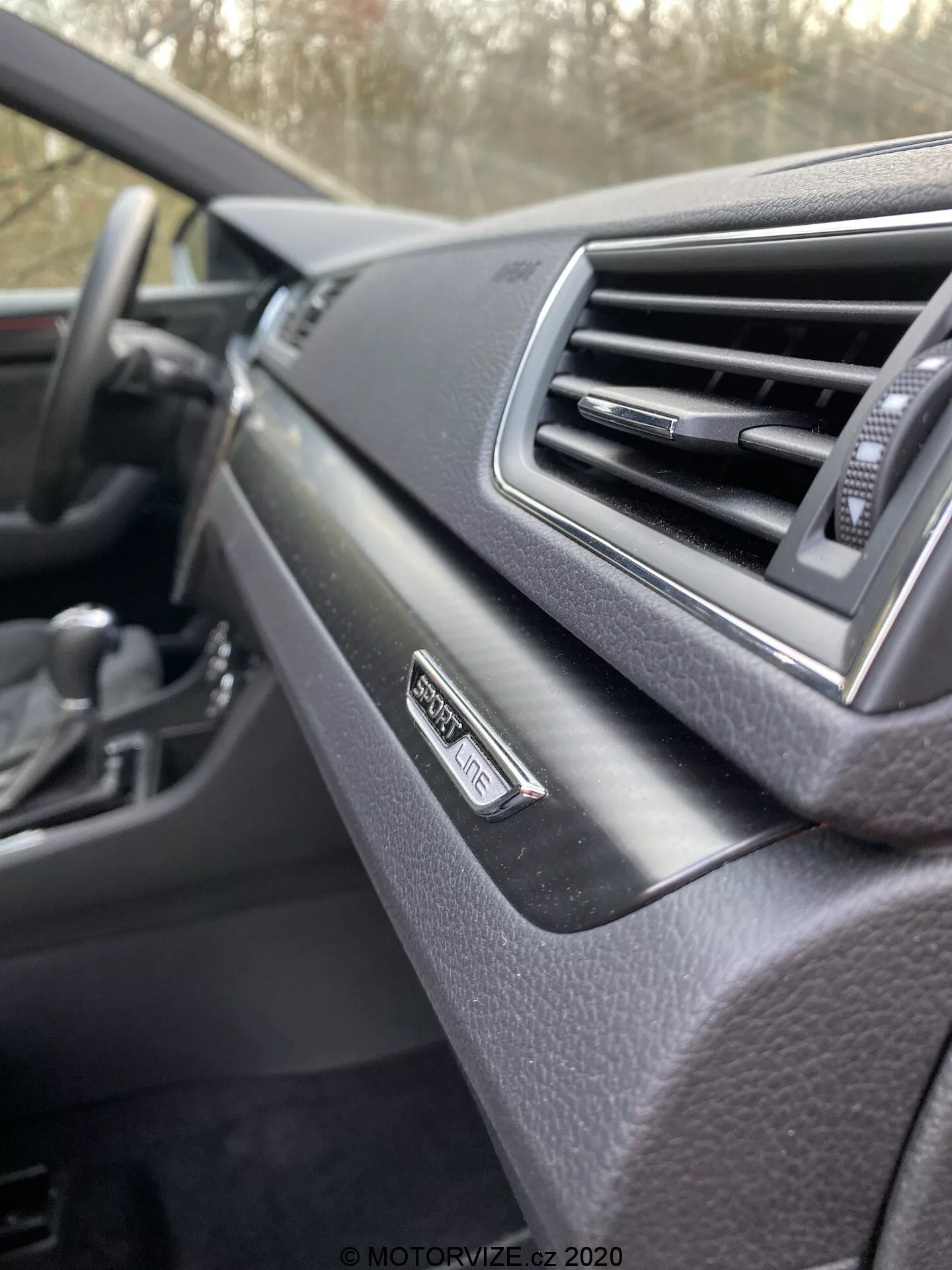 Cerca del interior del Škoda Superb 2019, salpicadero desde la perspectiva del pasajero. Enfoque en el emblema 'Superb' cerca de la ventilación, diseño moderno con acabado suave. Volante desenfocado en el fondo, esquema de color oscuro.