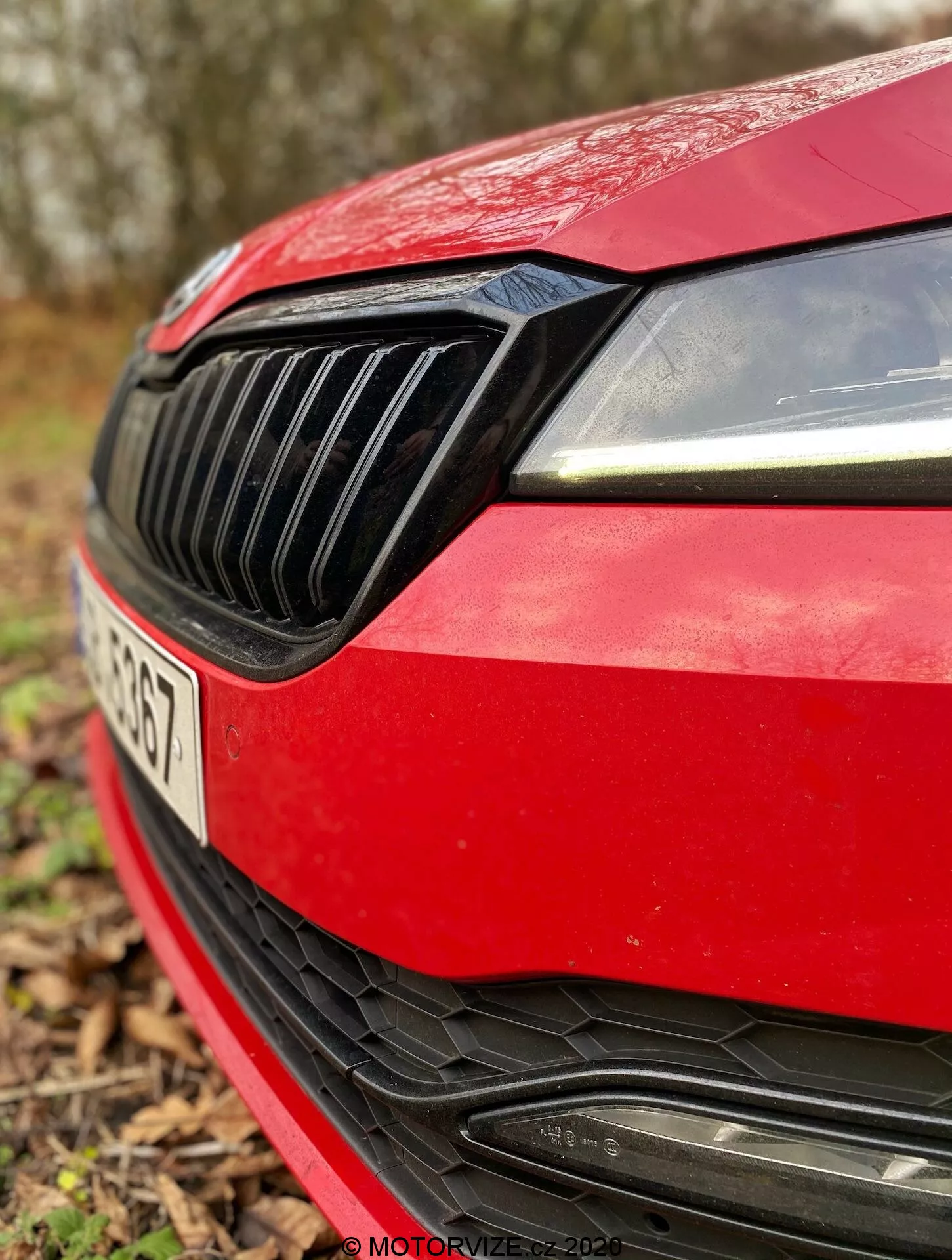 Vista de cerca del cuarto frontal izquierdo de un Skoda Superb Limousine 2019 rojo. La imagen captura el diseño moderno del faro derecho con luces diurnas LED, la parrilla negra sólida con listones verticales, el borde aerodinámico del capó rojo, el parachoques delantero deportivo con una gran entrada de aire con listones horizontales y el espacio para el emblema del fabricante, que está parcialmente oculto. La matrícula es ligeramente visible pero está fuera de foco debido a la poca profundidad de campo, con un fondo exterior natural desenfocado.