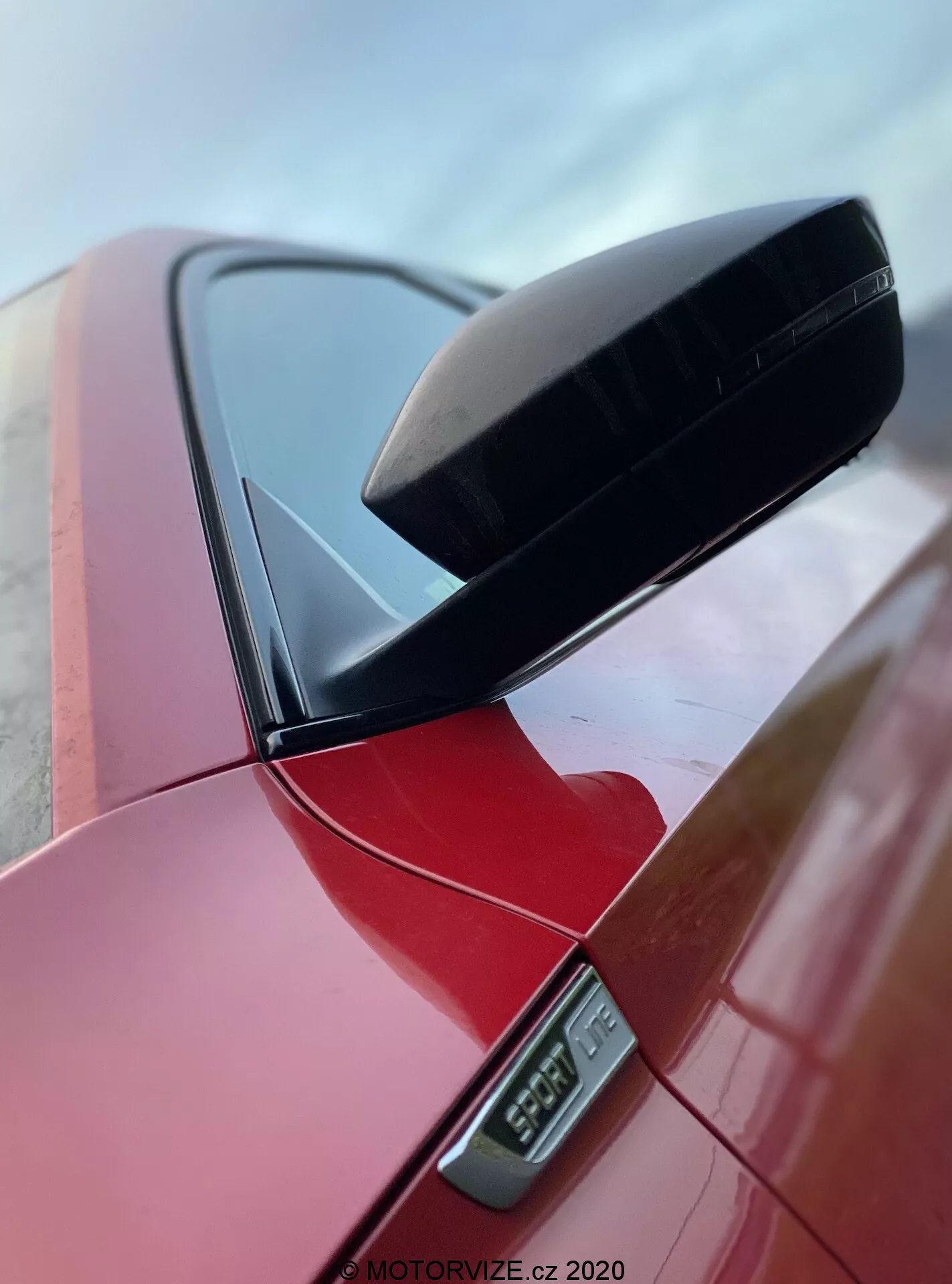 Vista de primer plano del espejo retrovisor lateral en un Skoda Superb 2019 berlina rojo vibrante, resaltando la carcasa aerodinámica negra del espejo y el indicador lateral LED debajo, con parte del pilar del coche visible, tomada desde un ángulo bajo que enfatiza el diseño limpio y el acabado pulido, marcada con una marca de agua MOTORVIZE.cz 2020.