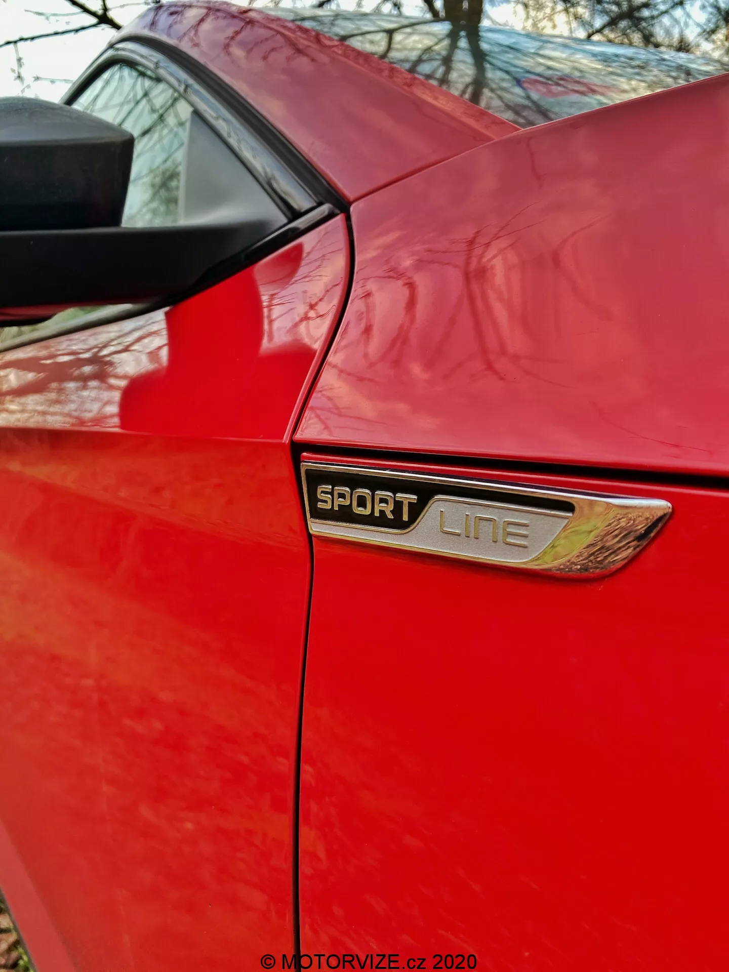 Vista en ángulo cercano del panel trasero de un Škoda Superb 2019 limusina rojo. Esta imagen se enfoca en el área detrás de la puerta lateral, destacando el emblema 'SPORT LINE' centrado en el cuadro con un borde cromado o metálico y un adorno de ventilación adicional debajo. El borde inferior de la ventana lateral trasera y el pilar trasero que sostiene la estructura del techo son visibles por encima del emblema. La foto resalta los niveles de equipamiento distintivos, características estéticas y calidad de la pintura de la edición Sport Line