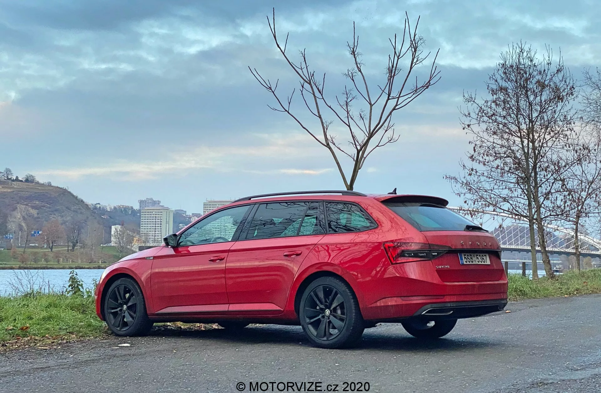 Imagen de un Skoda Superb 2019, station wagon, vista trasera de tres cuartos, mostrando luces traseras envolventes montadas en lo alto, parachoques robusto, sin tubos de escape visibles, spoiler en el techo, líneas de contorno laterales elegantes, manijas de las puertas del color de la carrocería, faldones laterales protectores, moldura de ventana cromada, ruedas de múltiples radios oscuras, estacionado en una superficie pavimentada cerca de la orilla del río con puente y árboles, cielo nublado, marca de agua MOTORVIZE.cz 2020.