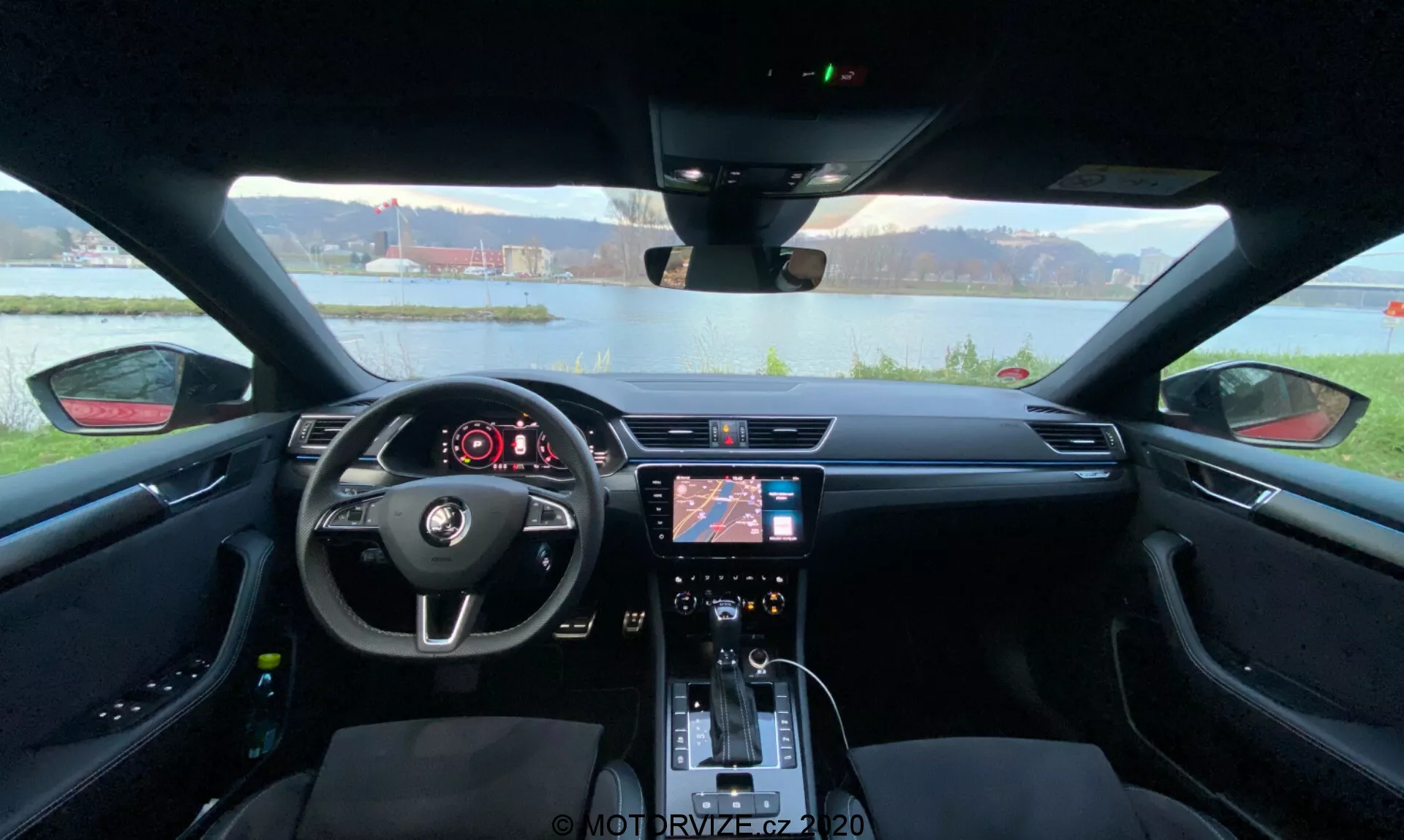 Vista interior de un Skoda Superb 2019 desde el asiento trasero mirando hacia el salpicadero, capturando todo el ancho del interior del coche. El volante central con controles integrados y el logotipo de Skoda es prominentemente visible en primer plano. Detrás del volante hay un panel de instrumentos digital que muestra información esencial del vehículo. La consola central cuenta con un sistema de infoentretenimiento con navegación y ajustes de medios, junto con controles de clima y botones de funciones debajo. La palanca de cambios, los portavasos y los asientos detallados con costuras de contraste destacan la estética moderna y se centran en la comodidad y conveniencia del conductor.
