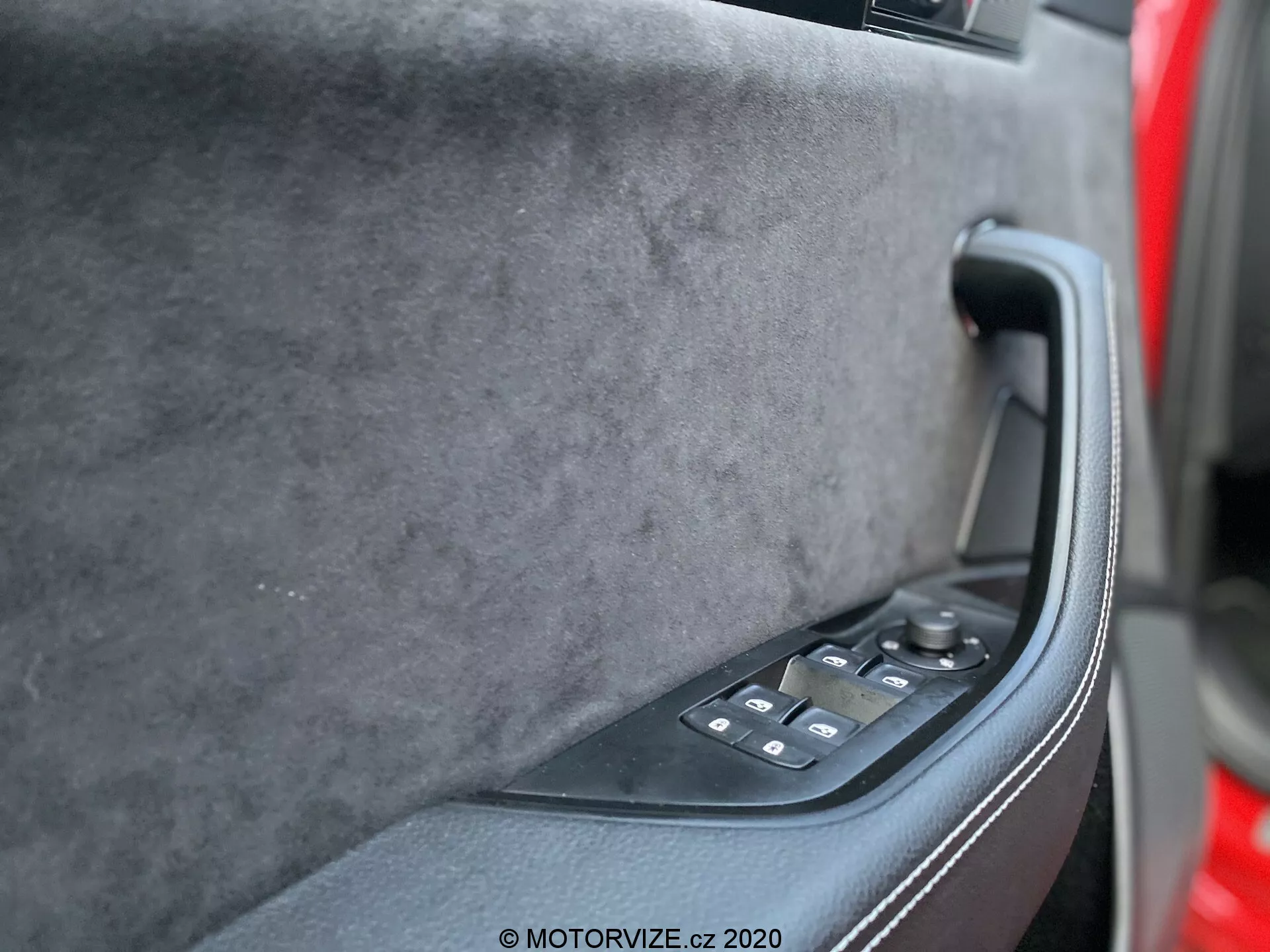 El panel interior de la puerta del Skoda Superb 2019 desde la perspectiva del asiento del conductor, mostrando la sección superior de tacto suave en tela premium, una manija ergonómica negra sólida y un panel de control negro brillante con ajustes de ventanas eléctricas y posiblemente controles de espejos eléctricos. Tonos oscuros apagados, tecnología moderna y un diseño lujoso son evidentes.