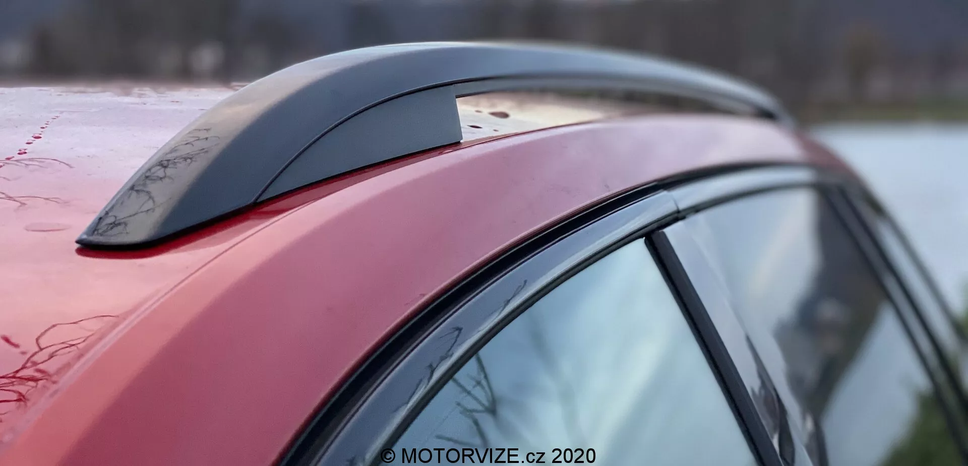 Vista lateral en primer plano de una sección del vehículo Škoda Superb 2019, enfocándose en la línea del techo rojo oscuro, el pilar lateral y el accesorio de antena tipo aleta de tiburón, con gotas de agua visibles que indican una superficie encerada o hidrofóbica; el diseño aerodinámico del automóvil es evidente a través de la curvatura y la pendiente del techo