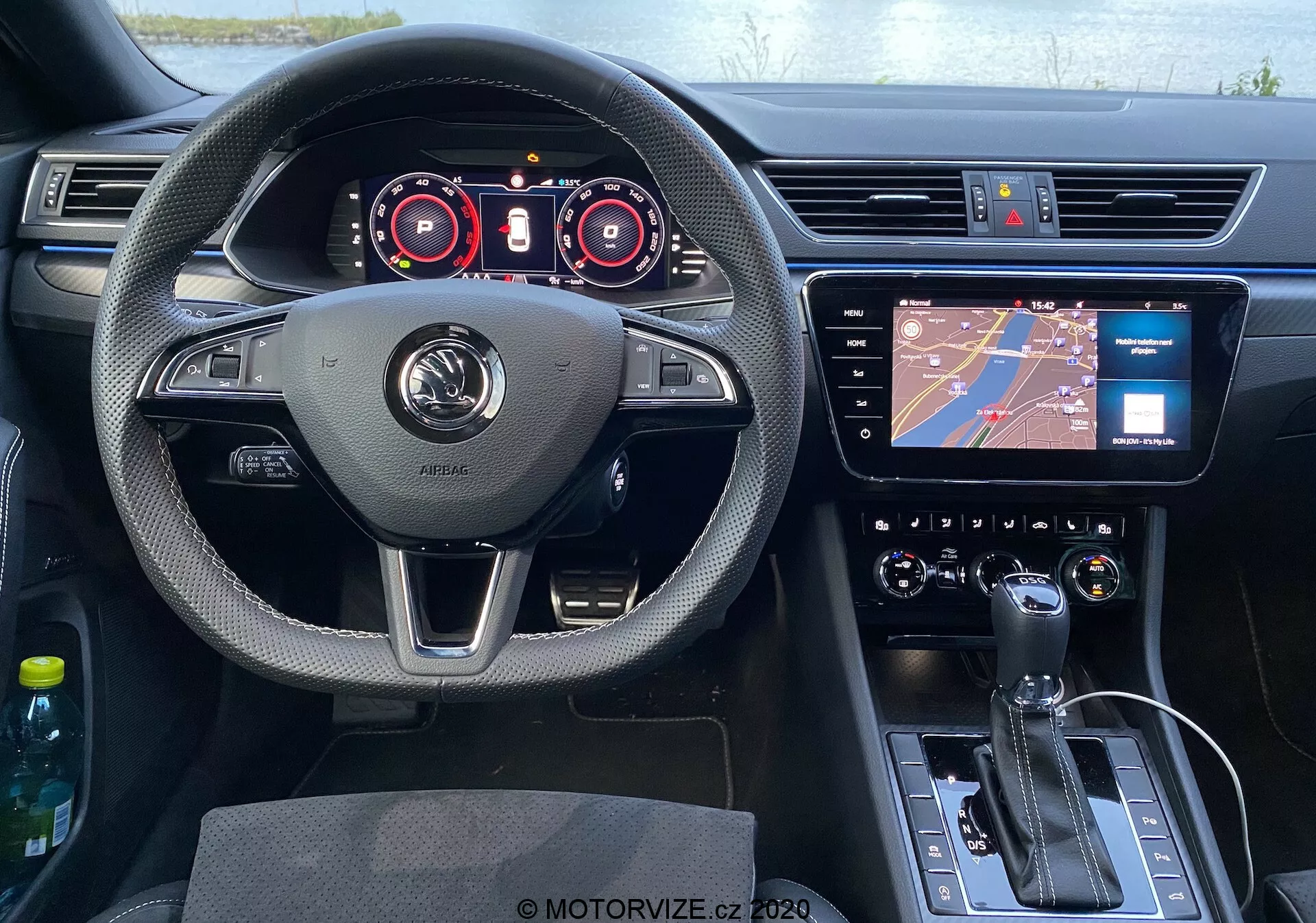 Vista del asiento del conductor del interior del Skoda Superb 2019 con un volante centralmente posicionado con soportes para los pulgares, botones multifunción y un destacado logotipo del fabricante. Detrás del volante hay un panel de instrumentos digital con indicadores para el velocímetro y tacómetro, junto con pantallas adicionales para detalles operativos. El centro del tablero alberga una pantalla táctil de infoentretenimiento con navegación, y debajo de ella, un sistema de control de clima con botones y diales. La consola central inferior incluye una palanca de cambios de transmisión automática, botón de freno de estacionamiento electrónico y un dial de control. El lado de la puerta del conductor y el espejo retrovisor son visibles, con controles evidentes de las puertas. La cabina presenta una mezcla de materiales texturizados, acentos cromados, volante forrado en cuero y plástico negro brillante en la consola, proporcionando una estética moderna.