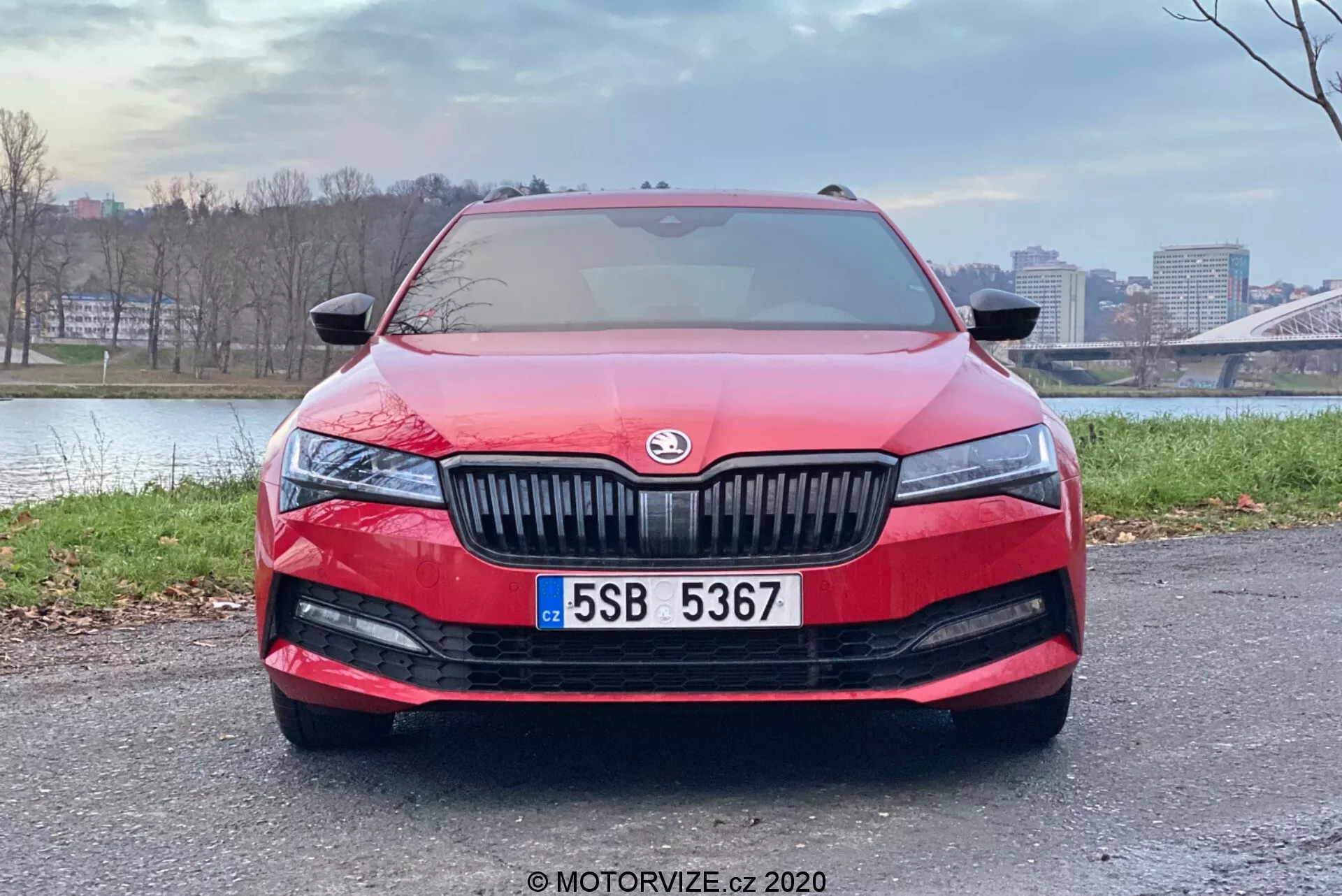 Vista frontal de un moderno Skoda Superb Limousine 2019 rojo, que muestra sus suaves contornos aerodinámicos, la prominente parrilla de láminas verticales con el emblema del fabricante en el centro, los estilizados y alargados conjuntos de faros LED, la amplia rejilla de admisión de aire en la parte inferior del parachoques con luces antiniebla, la matrícula central y posiblemente un sensor o cámara de sistemas avanzados de asistencia al conductor. El parabrisas refleja el cielo, sugiriendo sensores de lluvia para limpiaparabrisas o un sensor de luz para la activación automática de faros. El coche está aparcado en una zona pavimentada con un fondo urbano, destacando su diseño y características.