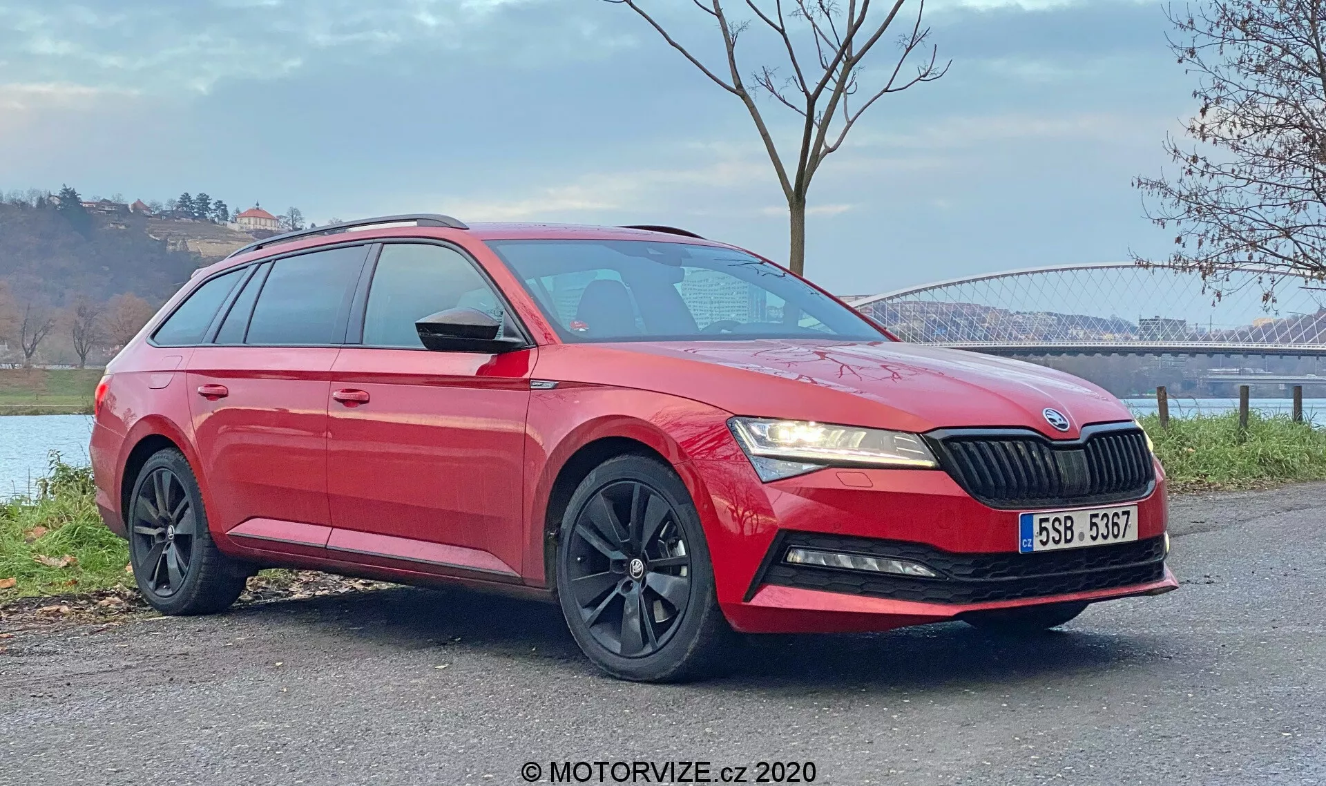 Imagen de un station wagon Skoda Superb 2019 rojo capturada desde una vista frontal de tres cuartos. La foto destaca la parrilla delantera con láminas verticales, faros angulares elegantes, luces antiniebla en el parachoques inferior, llantas de aleación negras de múltiples radios y carrocería brillante con líneas de carácter distintivas y rieles en el techo. También son visibles los espejos laterales color carrocería con señales de giro integradas y los cristales traseros tintados para privacidad. El vehículo, ubicado sobre una superficie plana al aire libre, se ve con un fondo natural que presenta un puente y un cuerpo de agua en la distancia. Las condiciones de luz diurna resaltan tanto las secciones inferiores como superiores del coche.