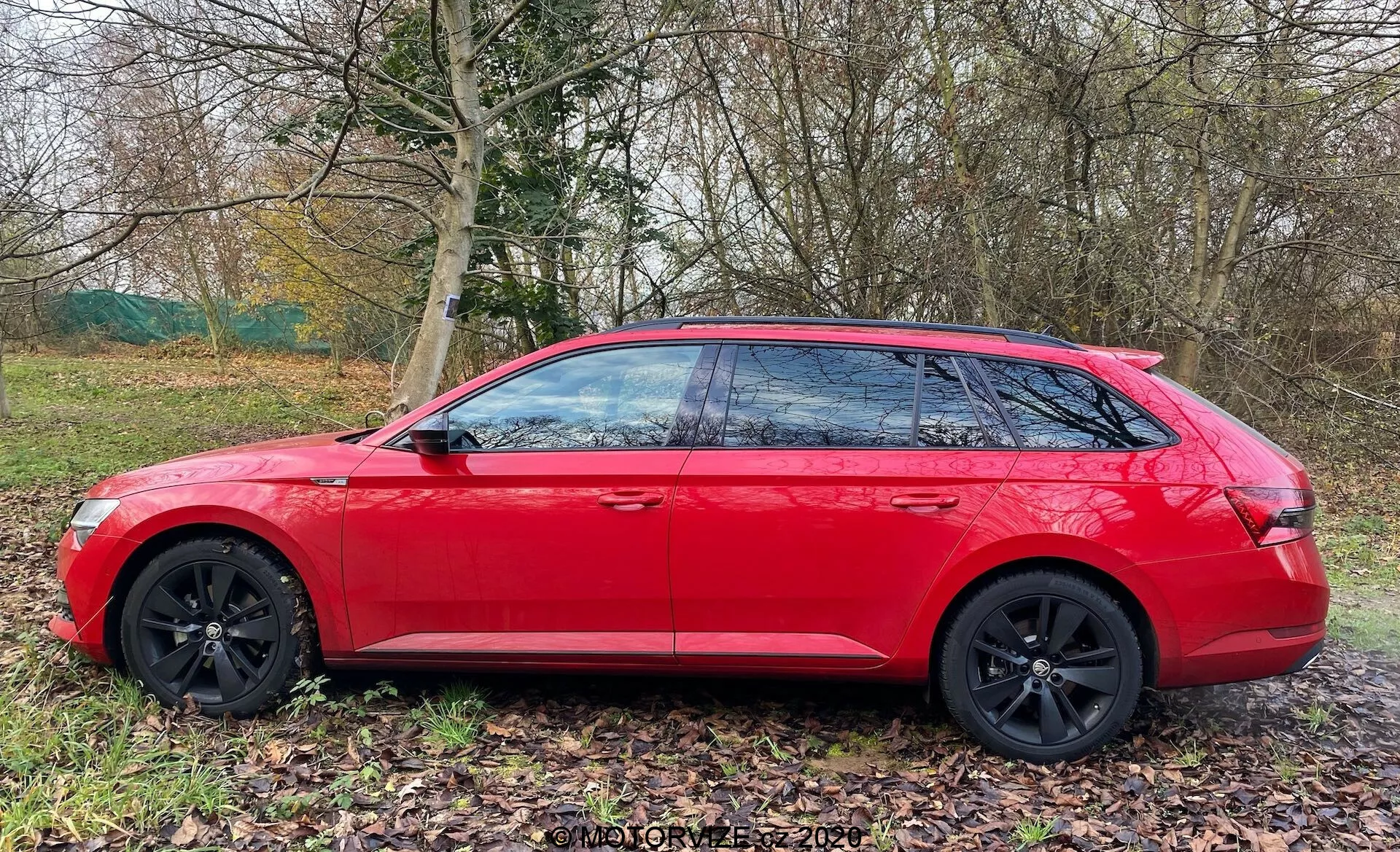 Station wagon Skoda Superb 2019 en rojo brillante, mostrado de perfil lateral. Características de diseño moderno que incluyen líneas geométricas nítidas, llantas de aleación negras de múltiples radios, ventanas tintadas con pilares B negros y una línea de techo inclinada para un perfil aerodinámico elegante. El coche está aparcado en una zona verde junto a un sendero, con naturaleza en el fondo, lo que sugiere que es adecuado para excursiones familiares o aventuras.