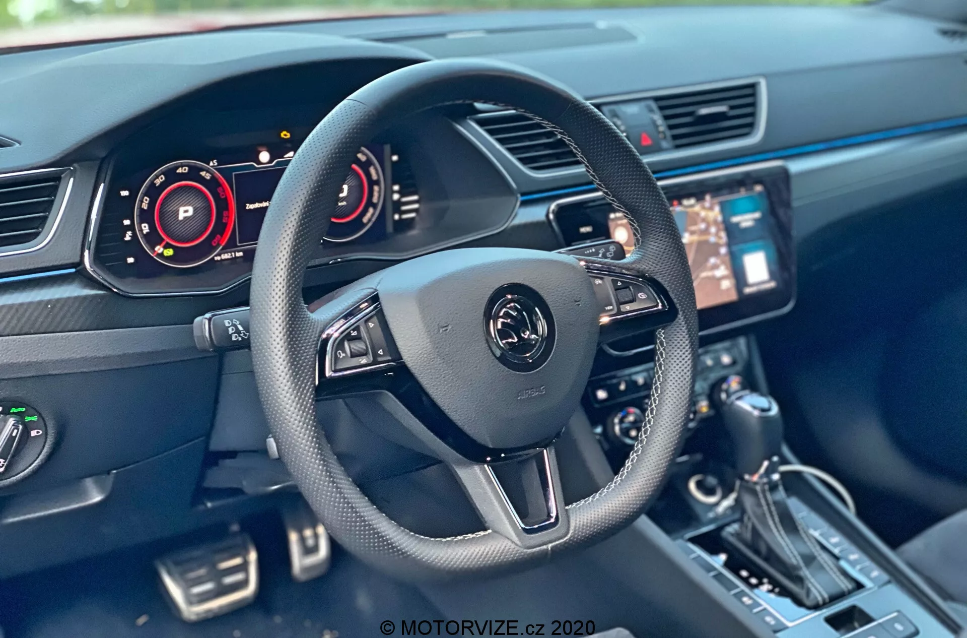 Interior del Skoda Superb 2019 desde la perspectiva del asiento del conductor, con un volante moderno de tres radios con controles multifuncionales y un emblema central. Componentes visibles incluyen un panel de instrumentos digital, sistema de infoentretenimiento central con pantalla táctil, consola, palanca de cambios, botones y sistema de control climático. La tecnología del interior del automóvil muestra elementos como tableros digitales y controles del volante.