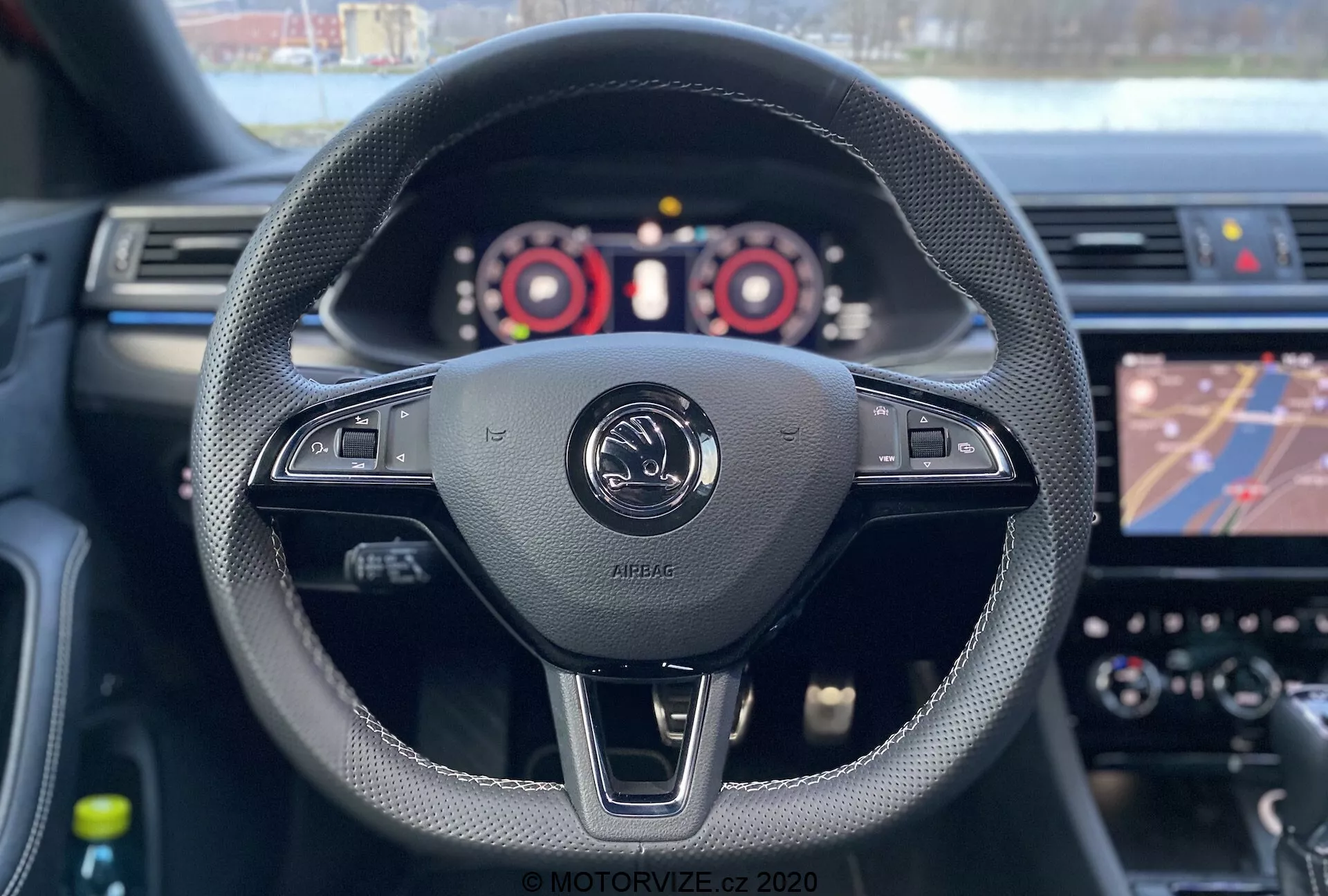 Vista interior del Skoda Superb Limousine 2019 desde la perspectiva del conductor. El asiento del conductor revela un volante envuelto en cuero negro con detalles cosidos, que alberga varios controles, incluidos los ajustes de volumen y posiblemente configuraciones del vehículo. En el centro de la imagen está el emblema del fabricante con la inscripción 'AIRBAG', destacando el airbag frontal del conductor. Se ven las paletas de cambio, lo que sugiere cambio de marchas manual. El panel de instrumentos con tacómetro, velocímetro y pantalla digital es parcialmente visible, mientras que el sistema de infoentretenimiento muestra un mapa de navegación. La poca profundidad de campo enfoca el volante y el emblema, con el tablero y la pantalla táctil ligeramente desenfocados.