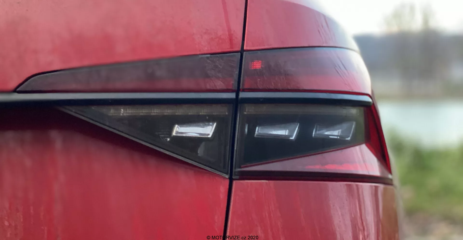 Sección en primer plano de la parte trasera del Skoda Superb 2019, enfocándose en el conjunto de luces traseras desde un ángulo lateral, mostrando un diseño moderno y elegante con elementos de luz LED horizontales encerrados en una lente de color rojo oscuro, reflejando una estética contemporánea deportiva o de lujo. La carrocería brillante y el trim negro horizontal añaden atractivo visual contra un fondo desenfocado.
