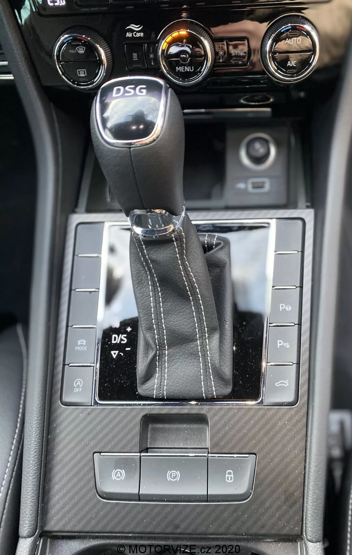 Vista interior de la consola central del Skoda Superb 2019 desde un ángulo hacia abajo, destacando la palanca de cambios DSG, botones de control simétricos, detalle texturizado similar a la fibra de carbono y controles de climatización con una pantalla táctil central que muestra información del vehículo.