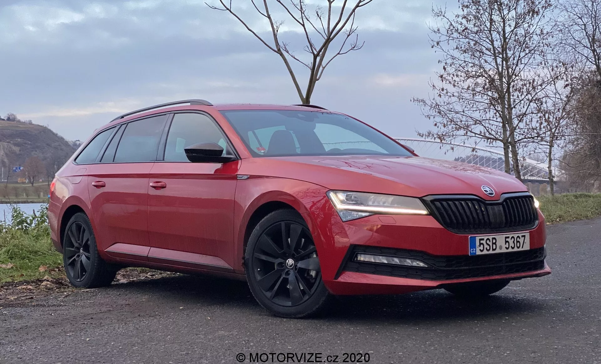 Imagen de un coche familiar Škoda Superb rojo en un ángulo de tres cuartos delantero, que muestra el diseño distintivo de la parrilla con el emblema de Škoda, faros LED, capó esculpido, luces antiniebla, llantas de aleación negras de varios radios, ventanas tintadas y espejos laterales con señales de giro integradas, situado en un entorno rural tranquilo con follaje y agua en el fondo.