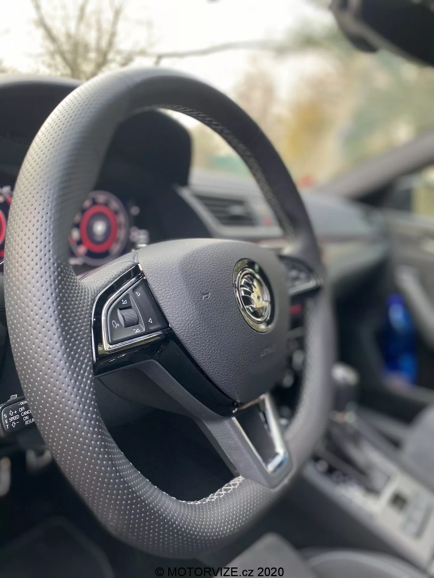 Vista interior de un Skoda Superb Limousine 2019 desde el asiento del conductor, enfocándose en un volante de fondo plano envuelto en cuero negro con controles multifuncionales. El logotipo del fabricante, con un emblema dorado sobre un fondo negro, se muestra prominentemente en el centro. Se puede ver el tablero y el cuadro de instrumentos detrás, destacando el diseño minimalista y de lujo del coche con diales contemporáneos detallados en rojo.