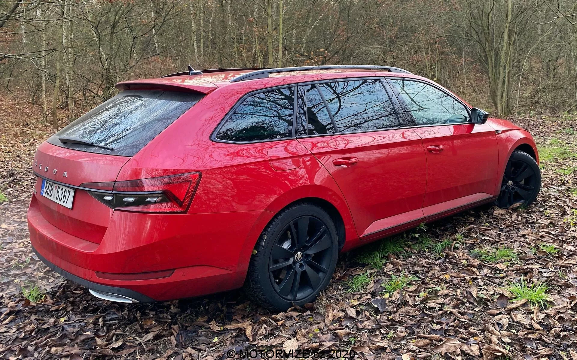Un station wagon rojo visto desde una vista trasera tres cuartos, posicionado fuera de la carretera sobre un lecho de hojas. Se captura el lado derecho del vehículo, desde el parachoques trasero hasta las puertas delanteras, mostrando su diseño moderno y premium. Las ruedas de aleación negras están ligeramente giradas hacia la derecha, agregando a su apariencia deportiva y de alta gama. Las características incluyen grupos de luces traseras elegantes, neumáticos de perfil bajo y un sistema de rieles en el techo. Las ventanas traseras tintadas sugieren mayor privacidad y protección solar.