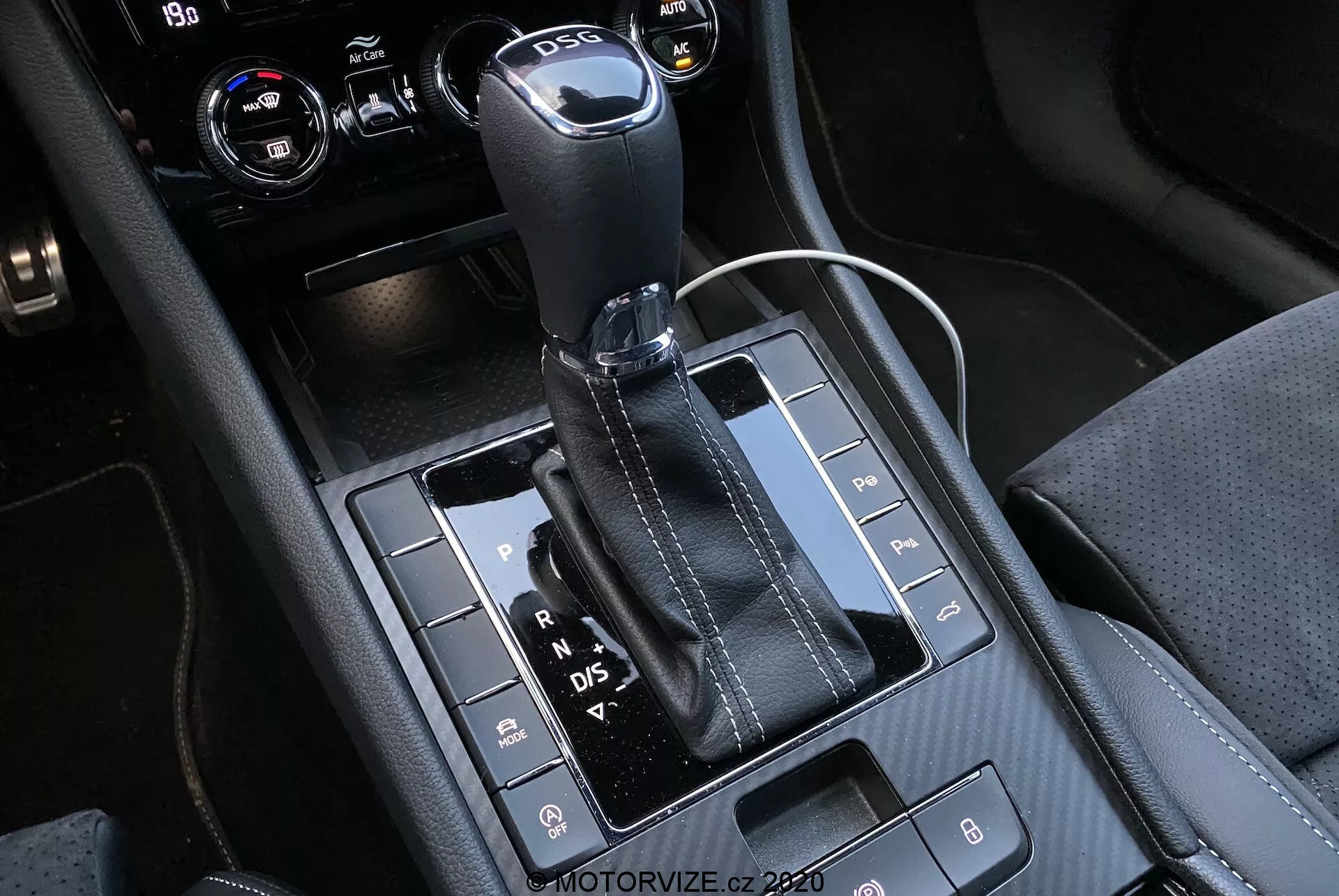 Vista de primer plano de la palanca de cambios automática y el área de la consola central del Skoda Superb 2019, con una palanca de cambios de cuero con parte superior metálica y letras visibles 'P, R, N, D/S' que indican diferentes marchas. El panel negro brillante alberga varios botones para funcionalidades como sensores de aparcamiento, desactivación del sistema Auto Start/Stop y control de tracción. La consola muestra una mezcla de materiales con costuras detalladas, incluyendo una toma de corriente de 12 voltios y posiblemente controles de climatización o de infoentretenimiento en la esquina superior derecha recortada.
