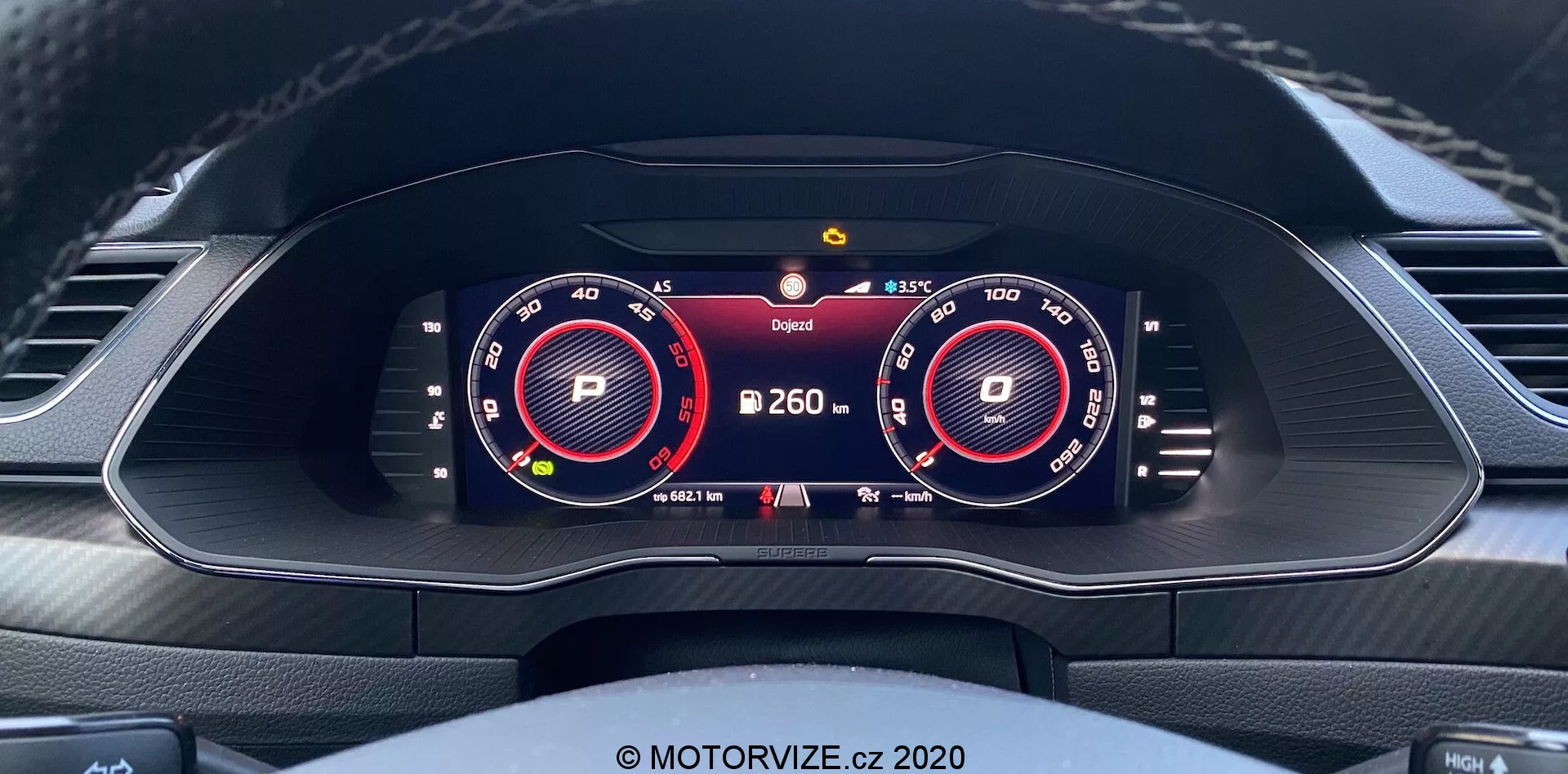Vista interior del Škoda Superb Limousine 2019 centrada en un conjunto de instrumentos totalmente digital y de alta resolución desde la perspectiva del conductor. La pantalla incluye dos grandes indicadores virtuales para el tacómetro y el velocímetro, iluminados en rojo y blanco. Entre los indicadores, se muestra información adicional como el indicador de marcha 'P', la lectura de temperatura y un símbolo de advertencia. Un indicador de batería sugiere capacidades eléctricas o híbridas. El diseño del salpicadero incluye salidas de aire y la parte superior de un volante contorneado con detalles de costura.