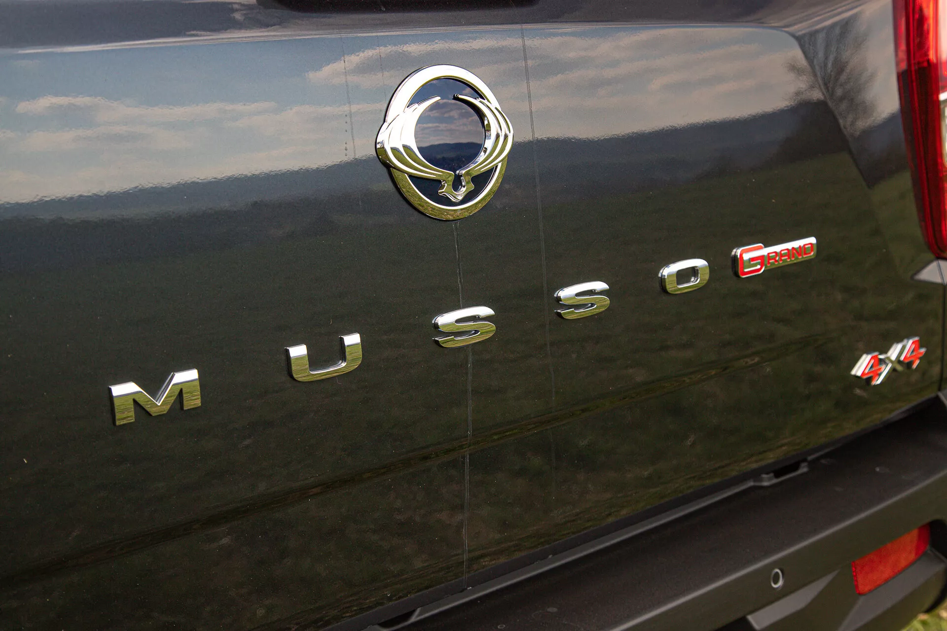 Vista detallada de la sección trasera de un SsangYong Musso Grand del 2022 mostrando el letrero cromado ‘MUSSO’, el emblema plateado del fabricante con un motivo estilizado de un pájaro, y la insignia roja ‘E-XDI Sport’ indicando el tipo de motor y nivel de rendimiento, configurado contra el exterior trasero oscuro, posiblemente negro, del vehículo con detalles cromados reflectantes y de textura.