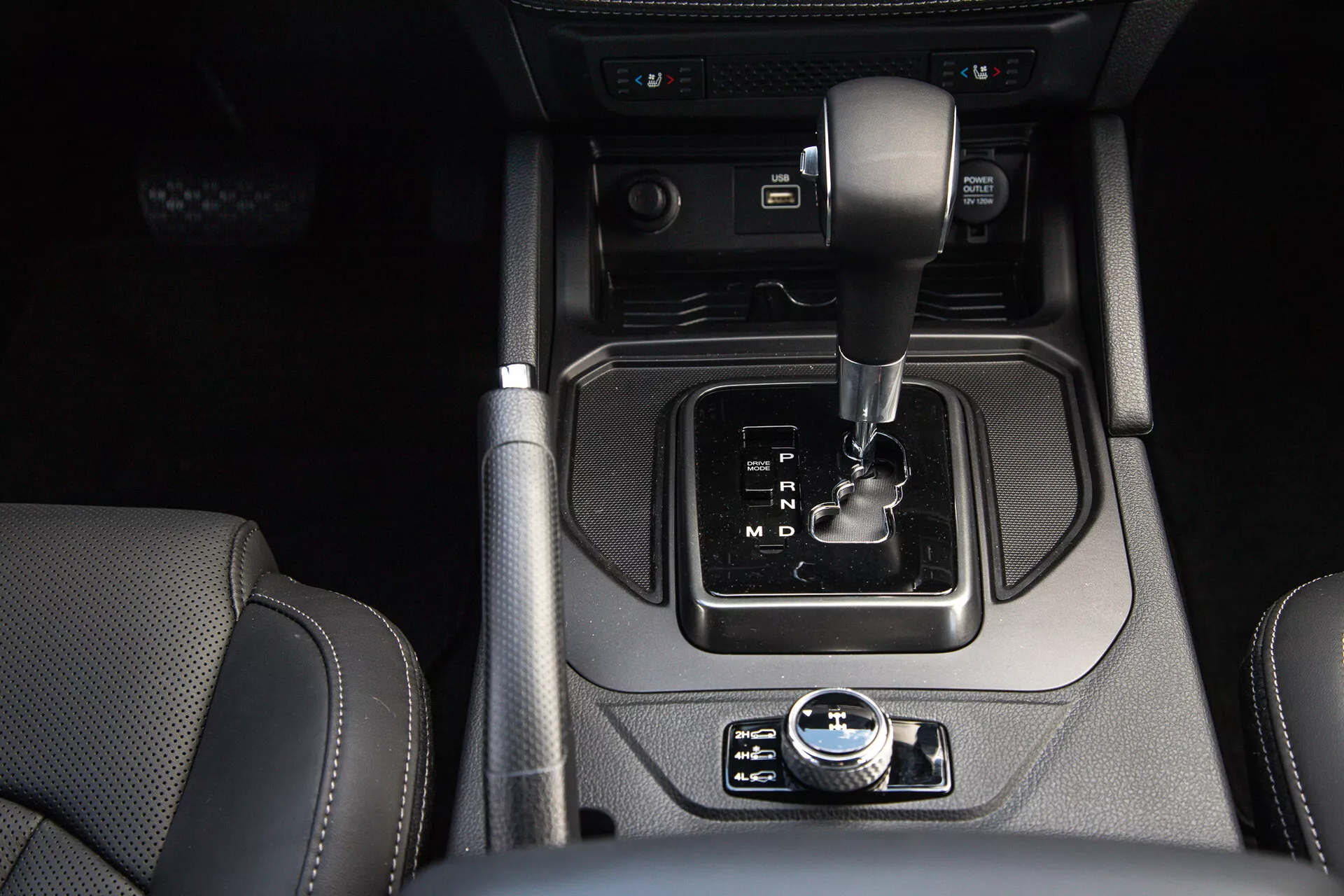 Vista detallada de la consola central del SsangYong Musso 2022 con palanca de cambios automática, freno de estacionamiento electrónico, botón de arranque/parada, interruptor del sistema de arranque y parada automático, portavasos, tomas de entrada auxiliar, y asientos revestidos en cuero visibles, destacando el diseño moderno y las características de lujo