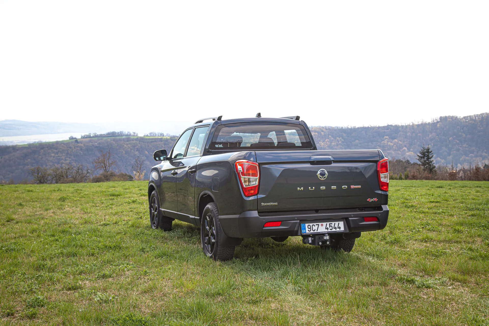 Camioneta SsangYong Musso Grand 2022 capturada desde un ángulo trasero destacando su apariencia deportiva y utilitaria, diseño de cuatro puertas y versatilidad al aire libre contra un escénico fondo de colinas y río durante el día