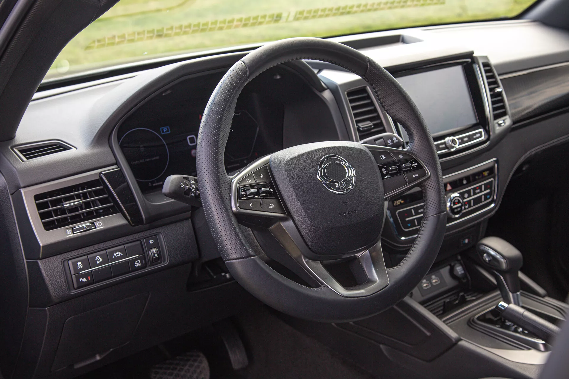 Foto del interior del SsangYong Musso 2022 mostrando el lado del conductor, incluyendo el volante multifunción de tres radios, el cuadro de instrumentos digital y el sistema de infoentretenimiento central en el tablero desde la perspectiva del pasajero trasero