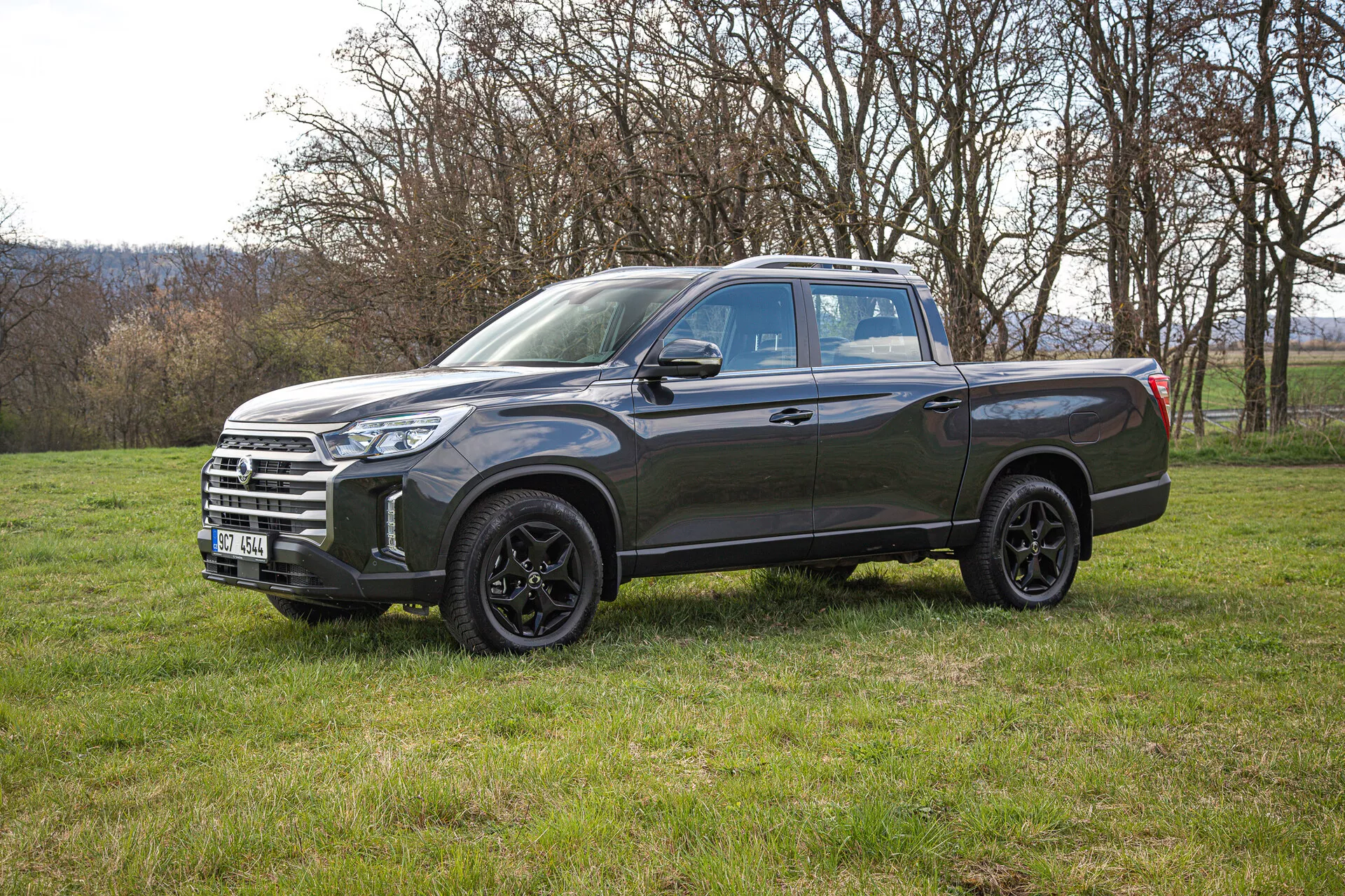 Vista lateral de una camioneta SsangYong Musso Grand 2022 de color gris oscuro sobre un campo de hierba, mostrando sus ruedas de aleación, faros LED y amplia caja de carga