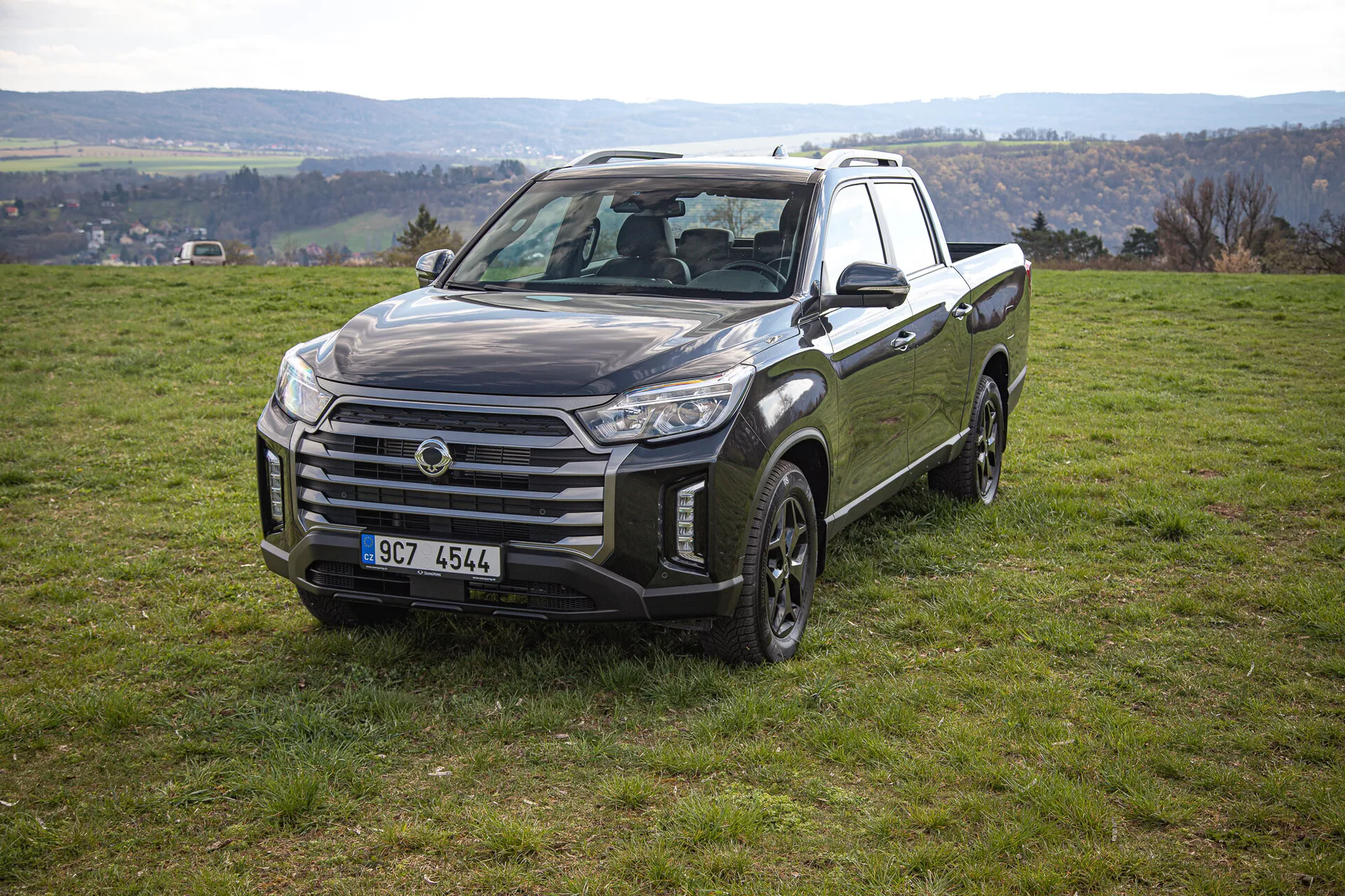Camioneta SsangYong Musso Grand 2022 en posición estacionaria sobre una inclinación con hierba mostrando vista frontal y lateral incluyendo parrilla grande, grupos de faros angulares y diseño de ruedas aptas para fuera del camino, con un paisaje rural de fondo.