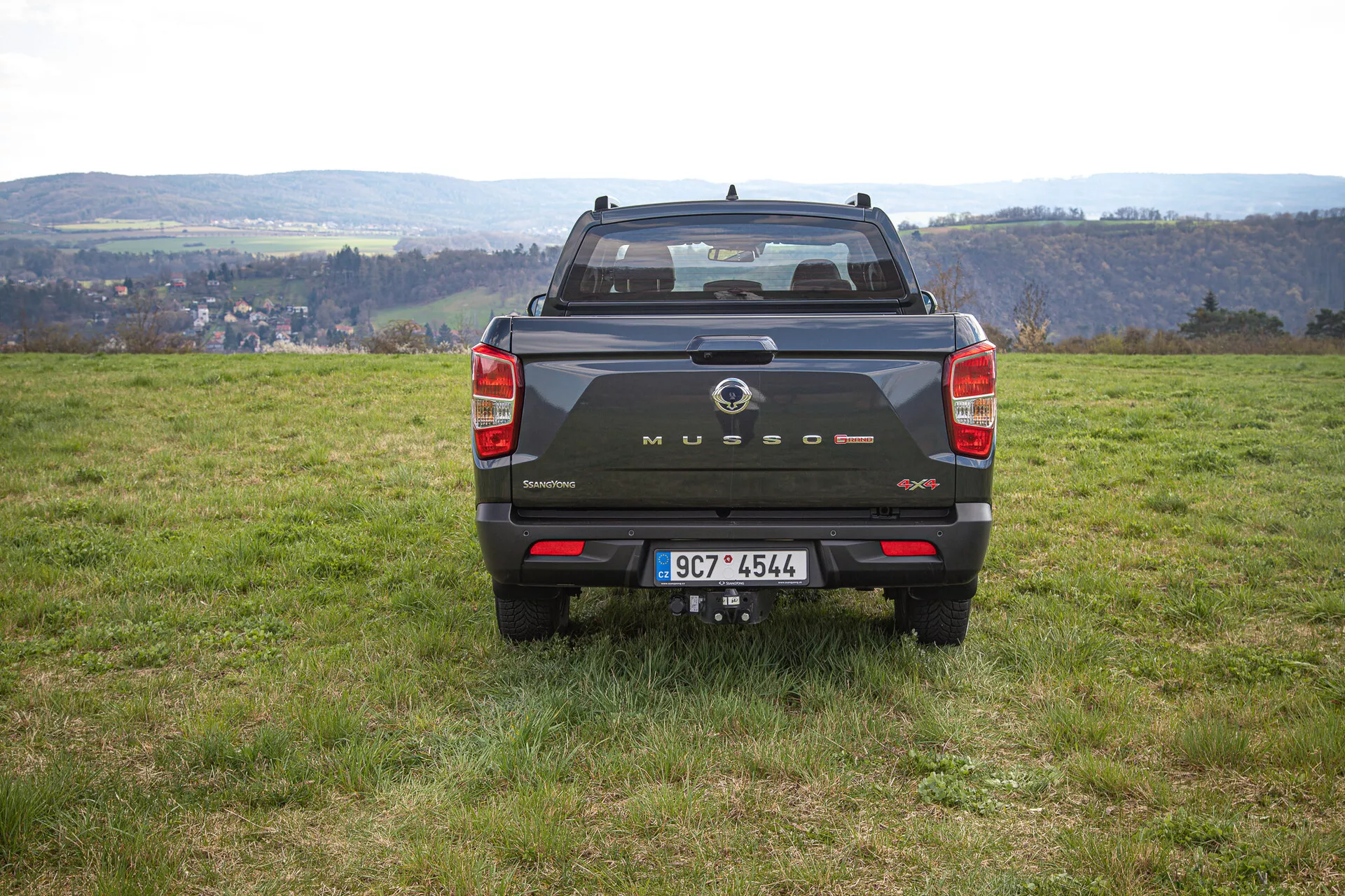 Vista trasera de una camioneta SsangYong Musso Grand 2022 de color oscuro sobre terreno irregular con césped con paisaje rural de fondo, mostrando la marca en el portón trasero, luces traseras verticales, sensores traseros y enganche de remolque durante el día