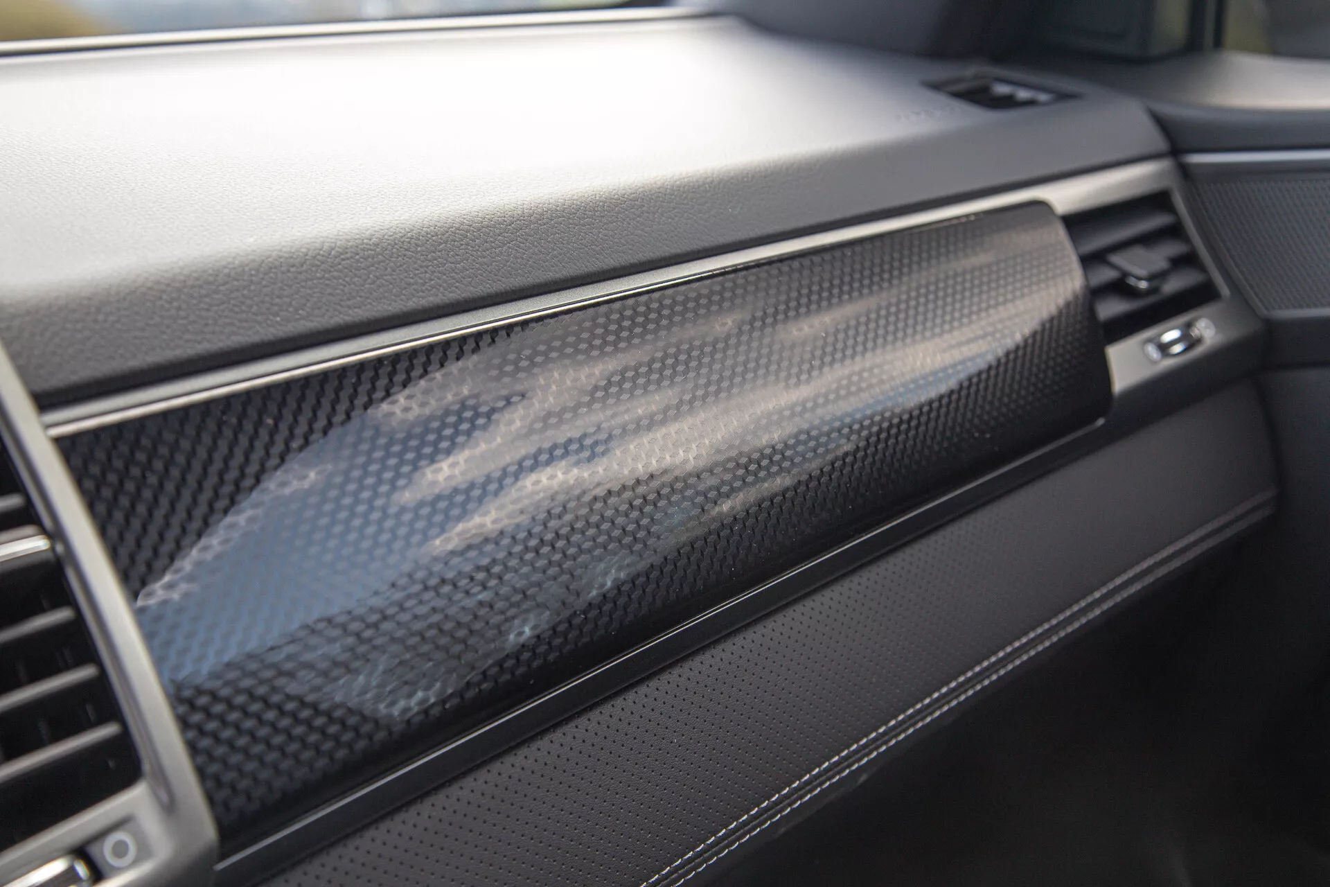 Vista de cerca del tablero del SsangYong Musso 2022 mostrando un lujoso acabado de fibra de carbono y materiales de alta calidad, enfocándose en el lado del pasajero con vislumbres de la guantera, ventilación y control de temperatura, ilustrando el énfasis del vehículo en el diseño interior moderno y la artesanía.