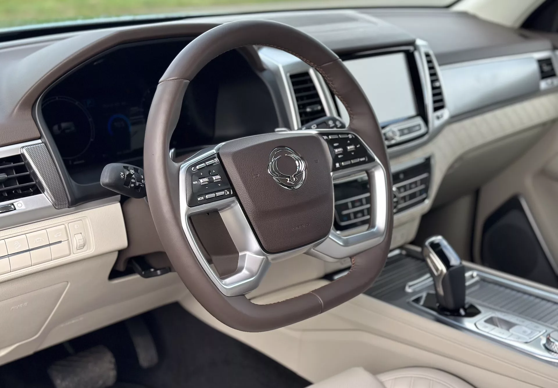 Vista interior lujosa del SsangYong Rexton 2021 desde el lado del copiloto, con volante multifuncional forrado en cuero con acentos metálicos, cuadro de instrumentos digital, sistema de infotenimiento avanzado y esquema de color en dos tonos beige y oscuro.