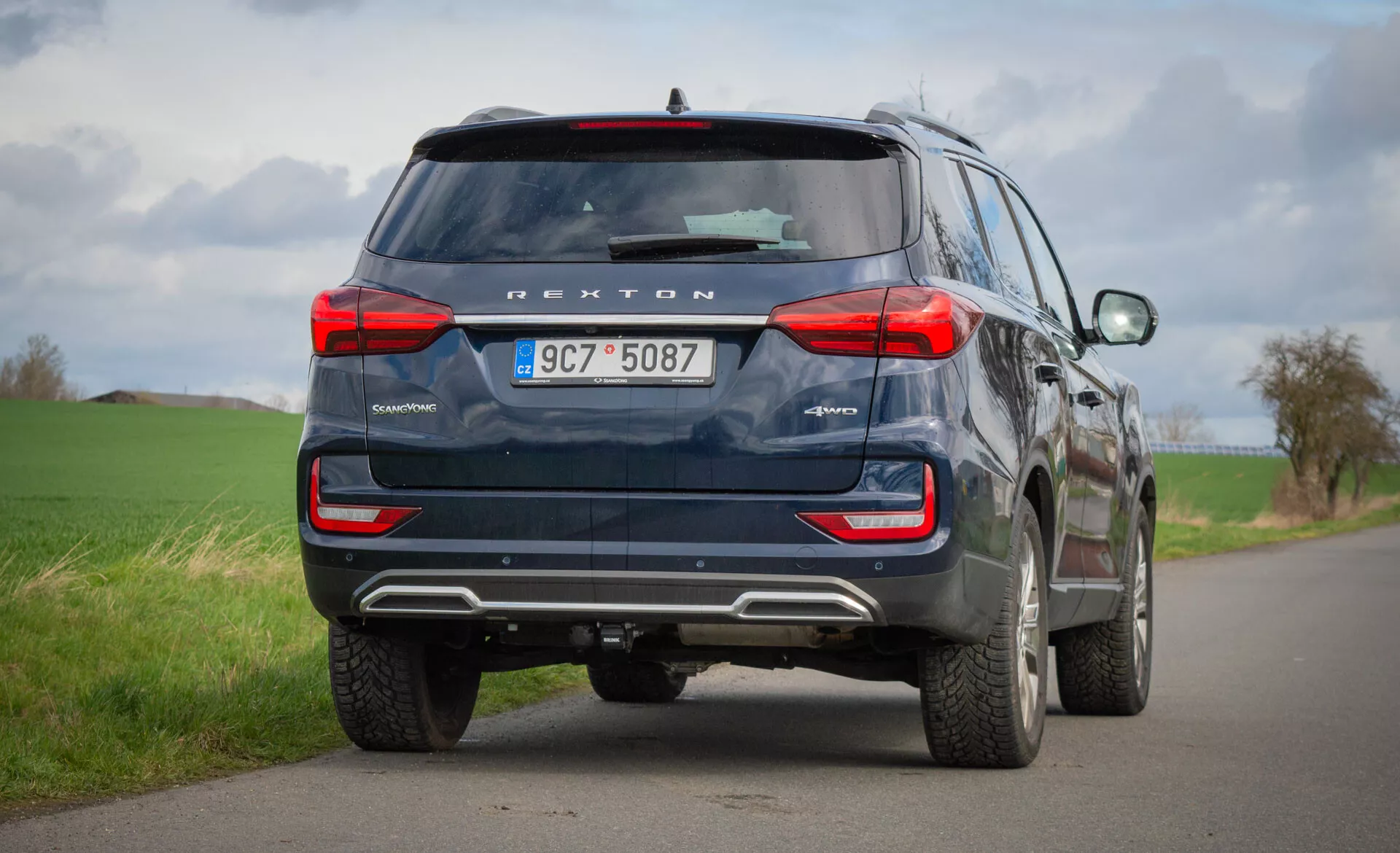 Vista trasera de un SUV azul SsangYong Rexton 2021 estacionado en un camino rural de dos carriles, destacando sus características de diseño como luces traseras, ventana trasera, tubos de escape y tecnología de seguridad, incluyendo cámara trasera y sensores de aparcamiento, bajo un cielo nublado.